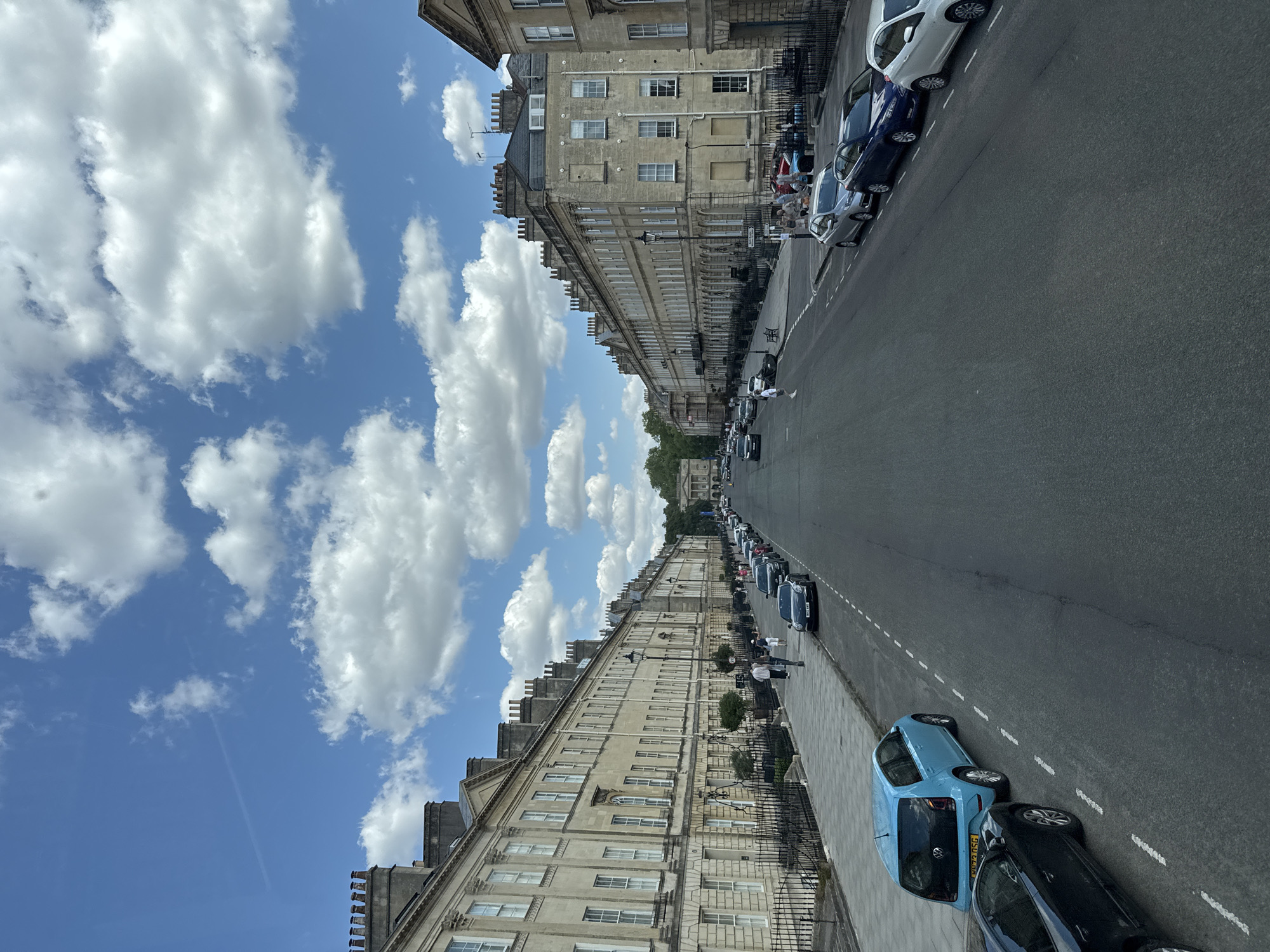 This is a street scene in Bath, United Kingdom, likely The Royal Crescent or a similar Georgian architectural ensemble. The view is down a wide, tree-lined street with imposing, pale stone terraced houses on both sides. The architecture is characterized by classical features, multiple stories, sash windows, and ornate chimneys. The street is paved, and there is a sidewalk with black metal railings in front of the houses. Cars are parked along the street, and a few people are walking on the sidewalk. The sky is blue with fluffy white clouds, suggesting fair weather. The overall impression is one of historical grandeur and a well-maintained urban environment.