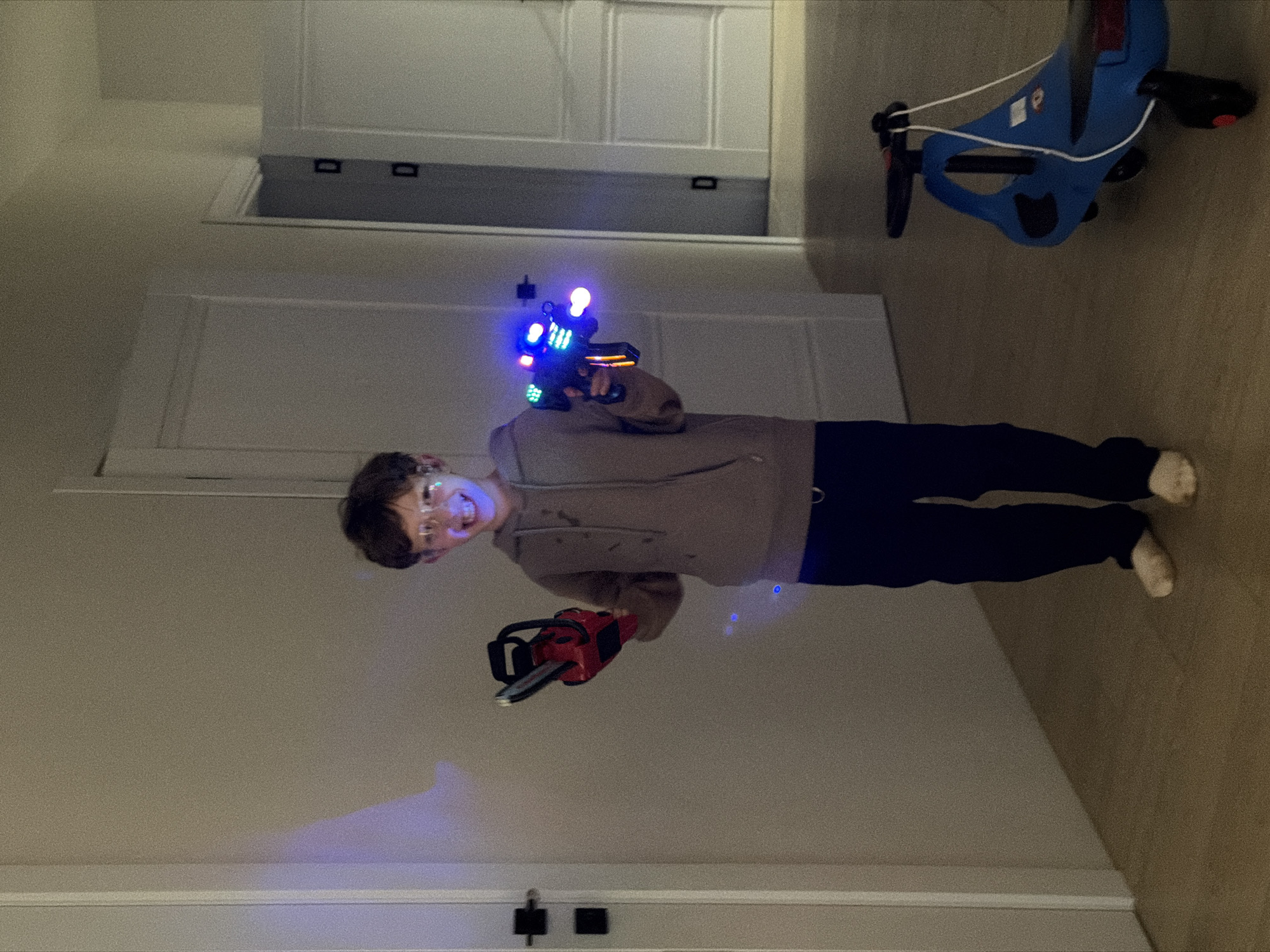 A young person, appearing to be a child or pre-teen, stands indoors and smiles broadly at the camera. They have short, dark hair and wear glasses, a light brown hoodie, and dark blue pants. In their right hand, they hold a toy laser gun with vibrant blue, purple, and green lights, which casts a faint blue glow on the light-colored wooden or laminate floor. In their left hand, they hold a red and black toy chainsaw.
The setting is an indoor room, likely a home, with plain light-colored walls and white paneled doors featuring black handles in the background. A blue children's ride-on toy with white straps is partially visible on the floor in the upper right. The lighting is artificial, characteristic of an indoor environment, making the toy lights particularly prominent. The subject's wide, toothy smile conveys joy and playfulness. No visible text is decipherable in the image. This media file was captured in Chaikovsky, Russia.