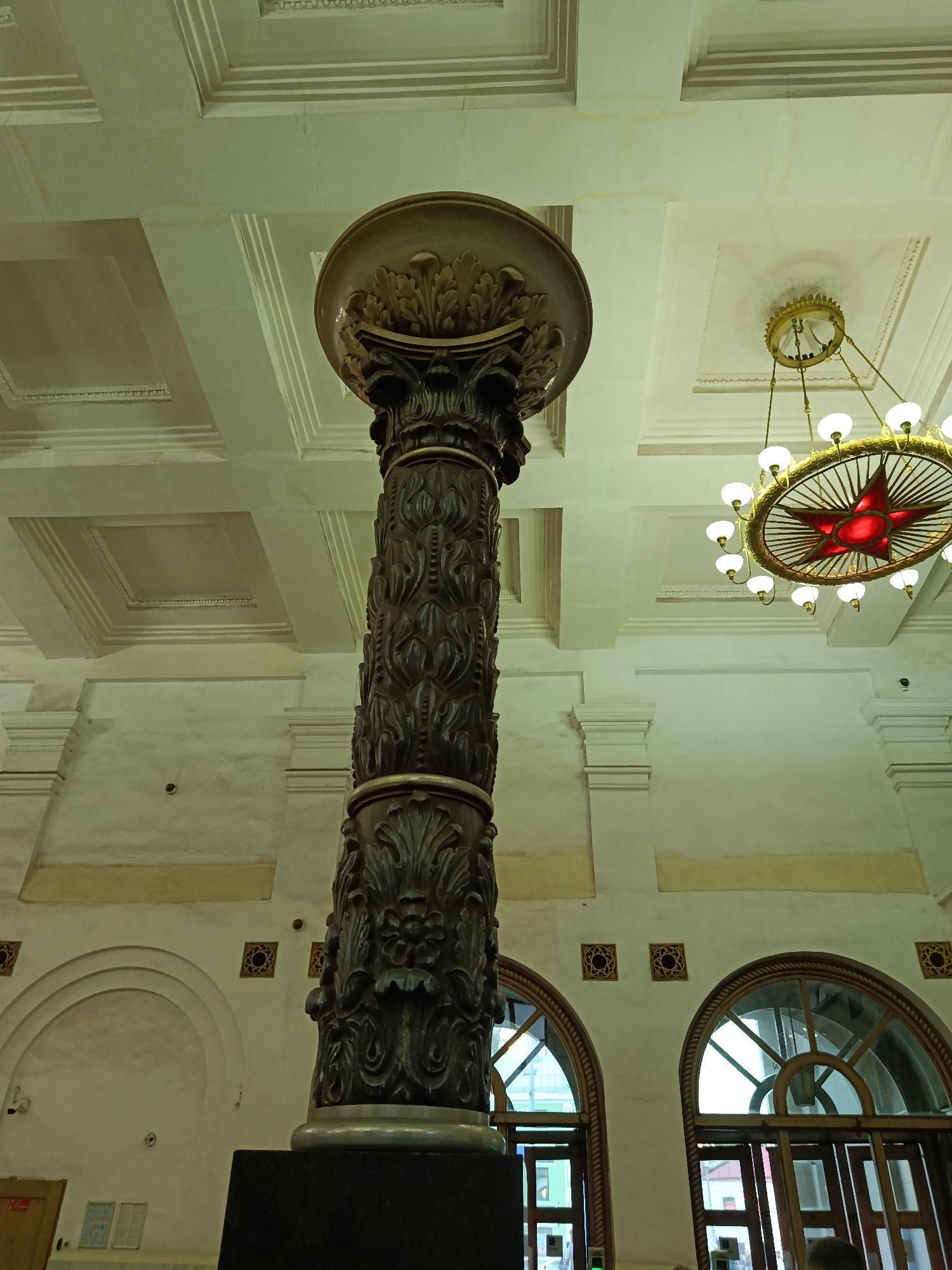 The image shows an interior view of a building with ornate architectural features. A tall, decorative column with intricate carvings dominates the foreground. The column is made of dark material and features floral and foliate motifs. The capital of the column is lighter in color and also decorated with leaves. Above the column, a multi-tiered ceiling with recessed panels and decorative molding is visible. A large, elaborate chandelier with multiple lights and a red star in the center hangs from the ceiling. The background shows large arched windows with wooden frames and panes, and walls with decorative elements and arches. The overall impression is of a grand and historic interior space, possibly a public building or a grand hall. The lighting is somewhat dim, with a greenish cast, suggesting artificial light sources. There are no visible people clearly depicted in the image. The location is identified as Unknown, Unknown.