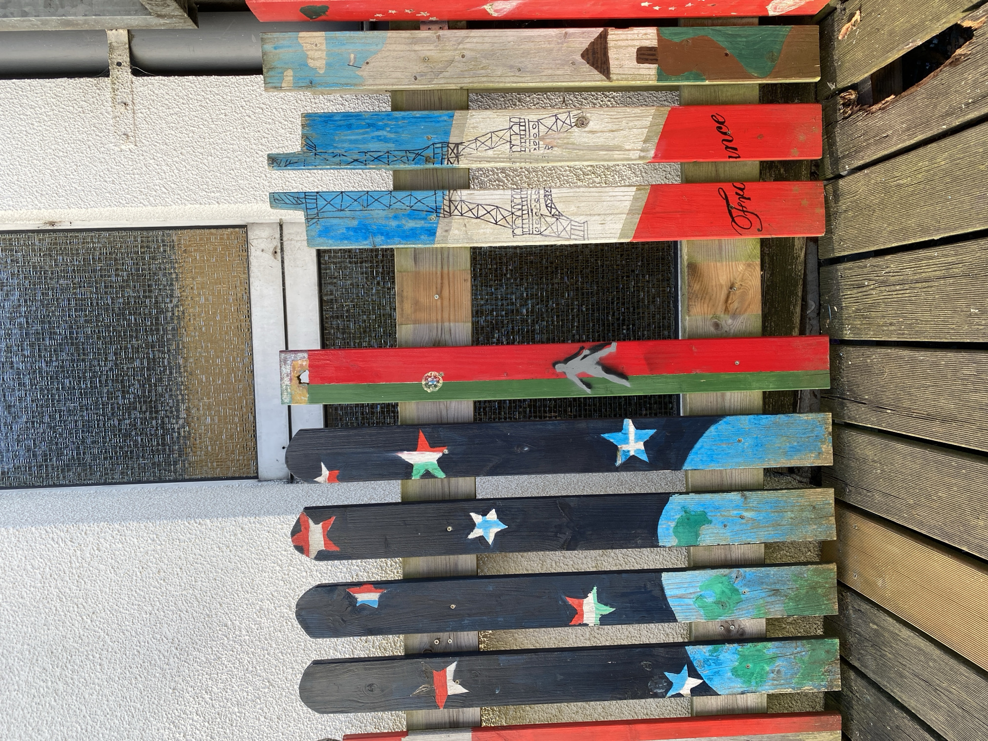 The image shows a close-up, eye-level view of a colorful, handmade fence made of wooden planks. The fence is installed in front of a textured white wall with a dark, crisscrossed window above it. The planks of the fence are individually painted with various designs, including stars, flags, and depictions of landmarks like the Eiffel Tower, suggesting a theme related to France and possibly other countries. Some planks are red, white, and blue, reminiscent of the French flag, with the word "France" painted in elegant script on two red planks. Other planks are black with white, blue, and red stars, as well as green and blue depictions of landscapes. The fence is supported by horizontal wooden beams. The ground in front of the fence is a wooden deck made of planks, some of which appear weathered and damaged. The lighting suggests it is daytime, with sunlight casting shadows. There are no people visible in the image. This fence is located in Altenkirchen, Germany, as it is a unique decorative feature associated with a specific private property within the city.