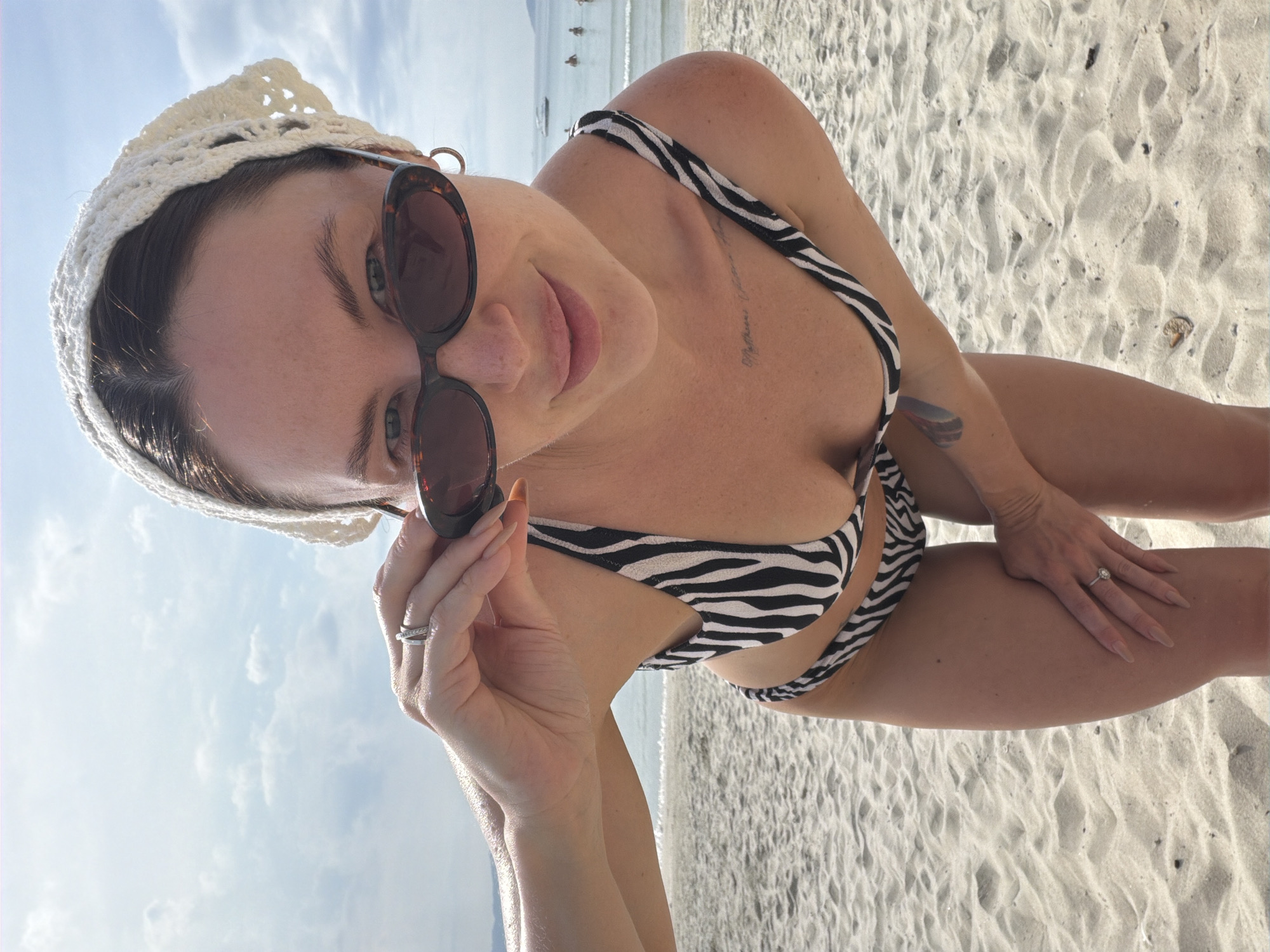 A woman wearing a white crochet headscarf and a zebra-print bikini bends over on a sandy beach, adjusting her dark-rimmed sunglasses. Her left hand is near her face, and her right hand rests on her thigh. A ring is visible on her left ring finger. In the background, the ocean meets a hazy sky with scattered clouds. A few small boats and figures are visible in the water. The location is specified as Dong Ninh Hoa Ward, Vietnam.