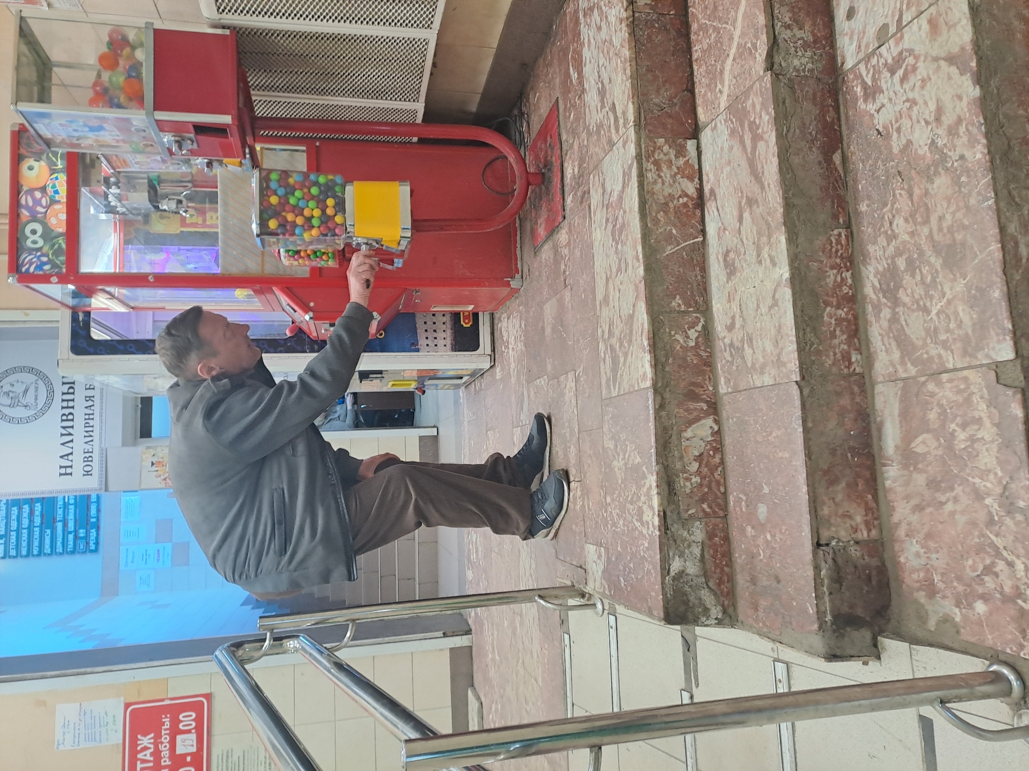 An adult male stands on the second step of a tiled staircase inside a commercial building in Minino, Russia, identified by Cyrillic signage indicating various retail sections such as clothing, textiles, and jewelry. He is positioned to the left of a group of vending machines and is operating a red and yellow gumball/candy machine with his right hand. The machine contains multi-colored spherical candies and stands on the tiled floor. Behind it is a larger red claw machine filled with toy capsules. The man is wearing a dark grey jacket, brown trousers, and dark athletic shoes. The staircase and surrounding floor are covered with reddish-brown and white marbled tiles, showing signs of wear and minor damage. A silver metal railing is visible to the left of the stairs. The background features light-colored walls, a white perforated panel, and various informational signs in Cyrillic.
