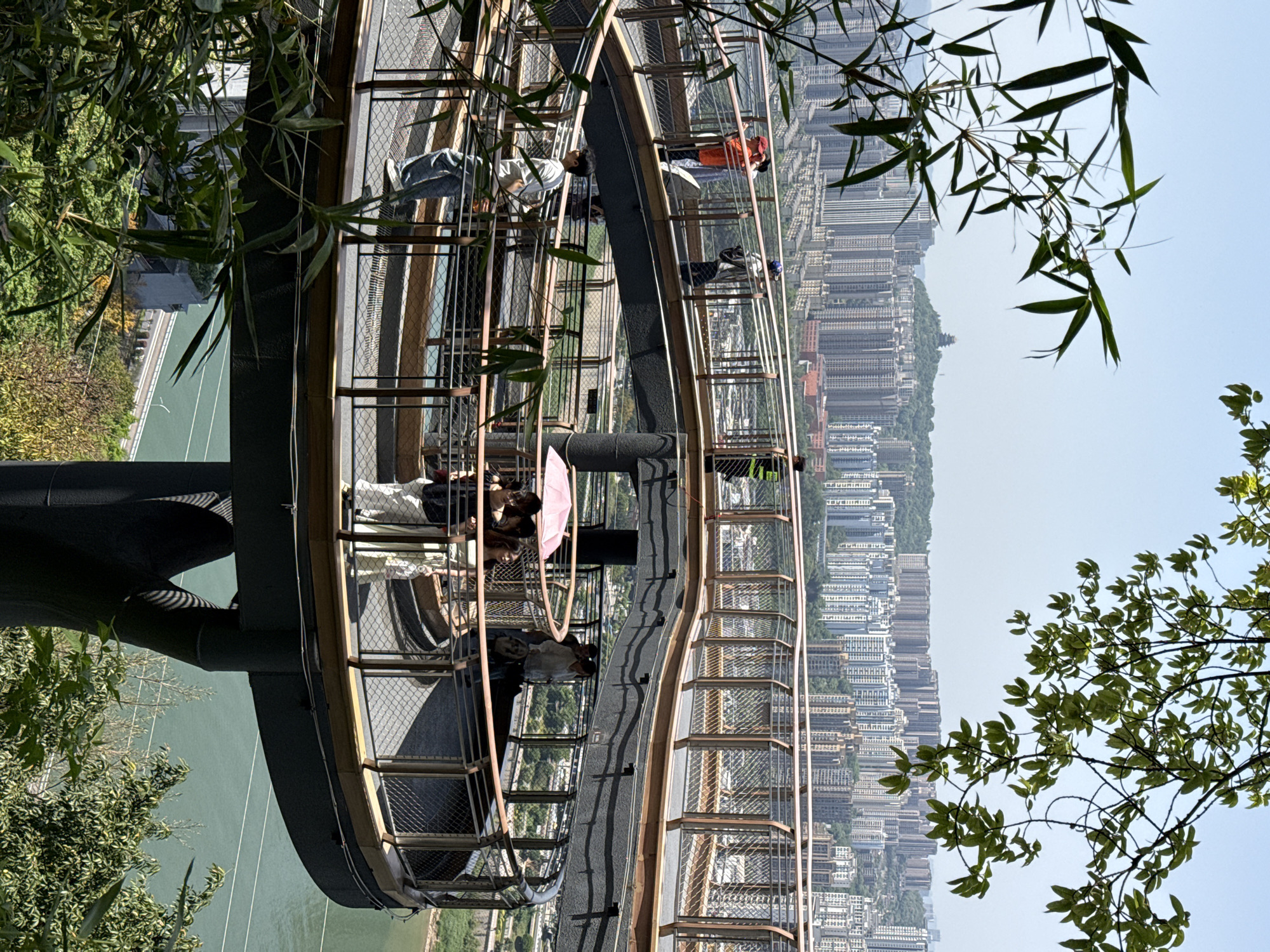 The image shows a high-angle view of a cantilevered steel and mesh walkway in the Yuzhong District of Chongqing, China. The walkway, populated with a few individuals, appears to extend outward from a hillside, offering panoramic views of the city and river below. The urban landscape is characterized by numerous tall buildings densely packed together. The sky is light blue, suggesting a clear day. Bamboo and tree branches frame parts of the view, adding a natural element to the urban setting. The architecture of the walkway includes metal railings and a dark-colored support structure. People are standing and observing the view from different points along the walkway.