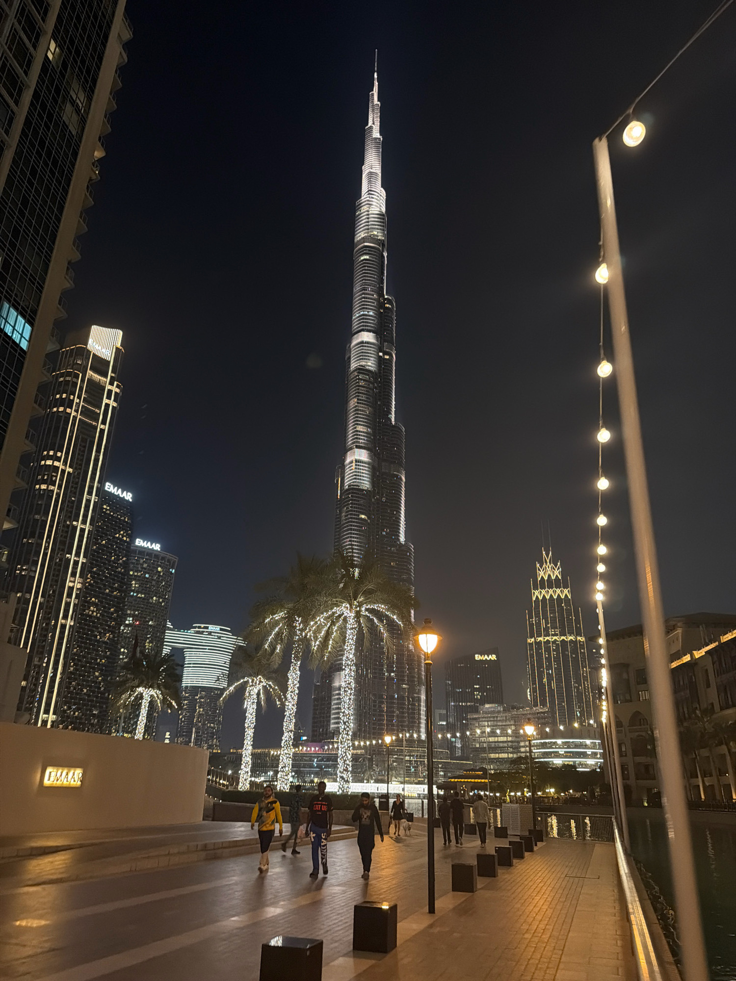 An eye-level, wide shot captures the Burj Khalifa skyscraper at night in Dubai, United Arab Emirates. The iconic tower is illuminated and dominates the background, with smaller, illuminated buildings and palm trees decorated with lights in the mid-ground. A paved walkway with square planters runs along the right side of the frame, and several people are walking on it. The walkway is lit by street lamps. A body of water is visible to the right of the walkway. The scene appears to be a public plaza or promenade.