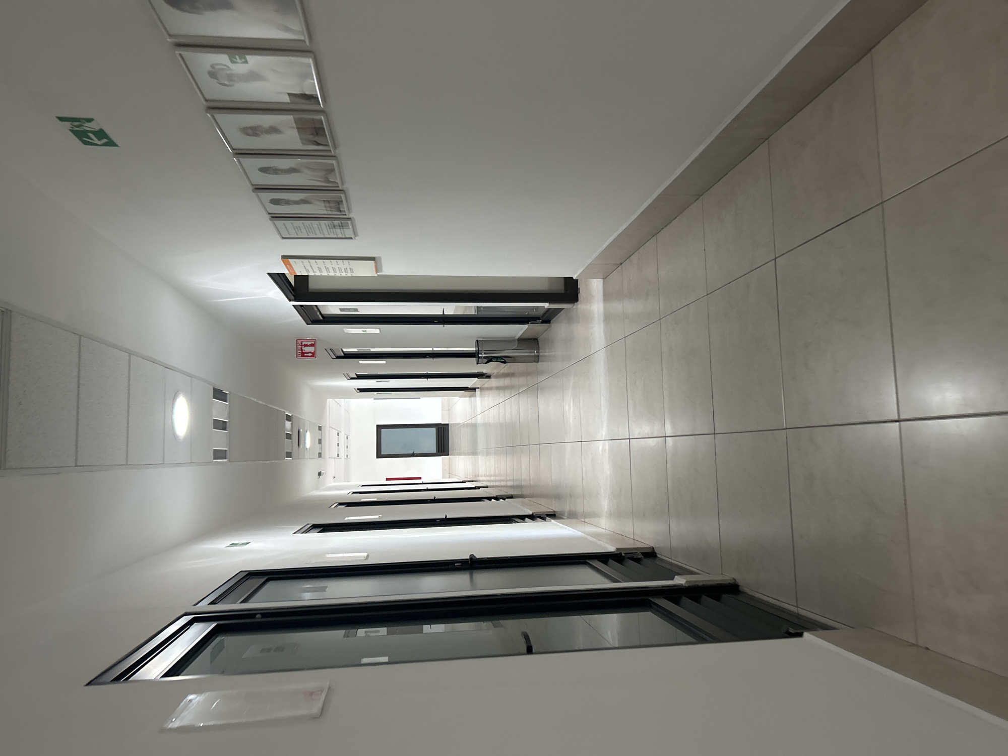 The image shows a long, well-lit hallway with a white ceiling and tiled walls. There are several framed pictures and a green exit sign on the ceiling at the left. Doors with black frames line the left side of the hallway, with windows in some of them. Towards the end of the hallway, there is a window. The location is an interior space, city and state are unknown. No people are visible in the image.