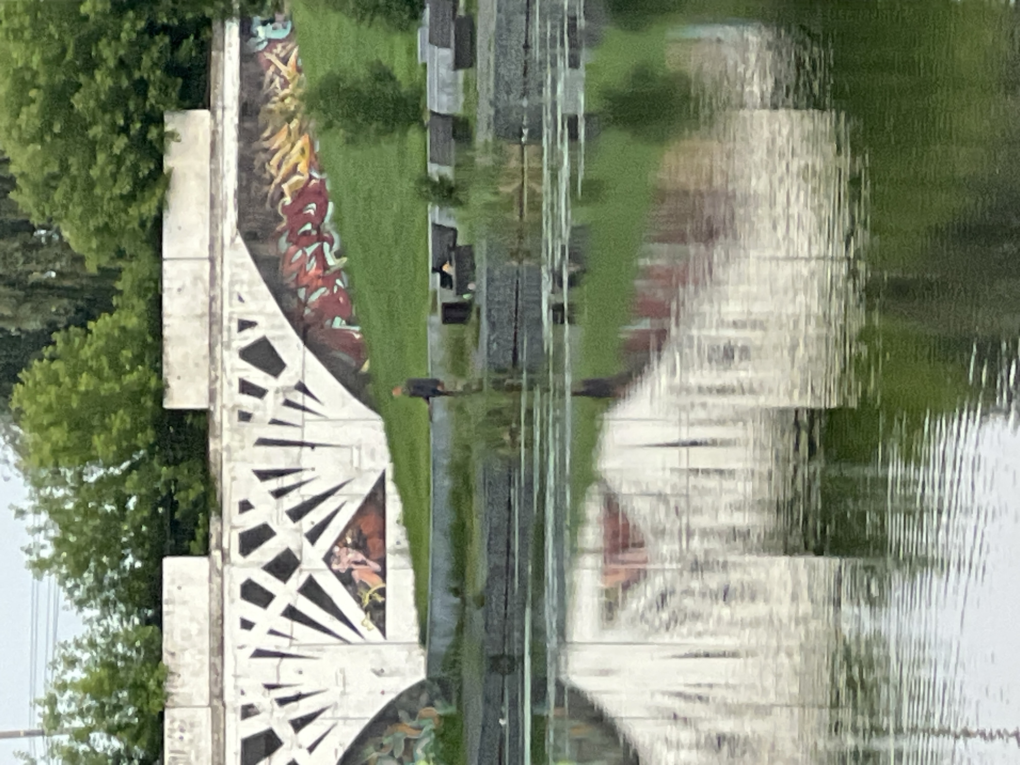 The image depicts a section of the embankment of the Pyshma River in Votkinsk, Russia, characterized by a concrete retaining wall adorned with graffiti. The wall features a geometric design with sharp, angular black shapes against a white background, interspersed with sections of colorful graffiti, including stylized text and imagery. Below the wall, a grassy slope leads down to the river, with several dark, rectangular benches positioned on the embankment.
In the middle ground, a lone individual stands on the embankment, facing away from the camera and appearing to be engaged in fishing, holding a fishing rod. The river in the foreground reflects the concrete wall and the surrounding greenery, creating a distorted, abstract pattern. The overall atmosphere is overcast, with a muted color palette dominated by greens, grays, and whites, characteristic of a cloudy day. The scene conveys a sense of urban recreation and artistic expression integrated into the natural environment.