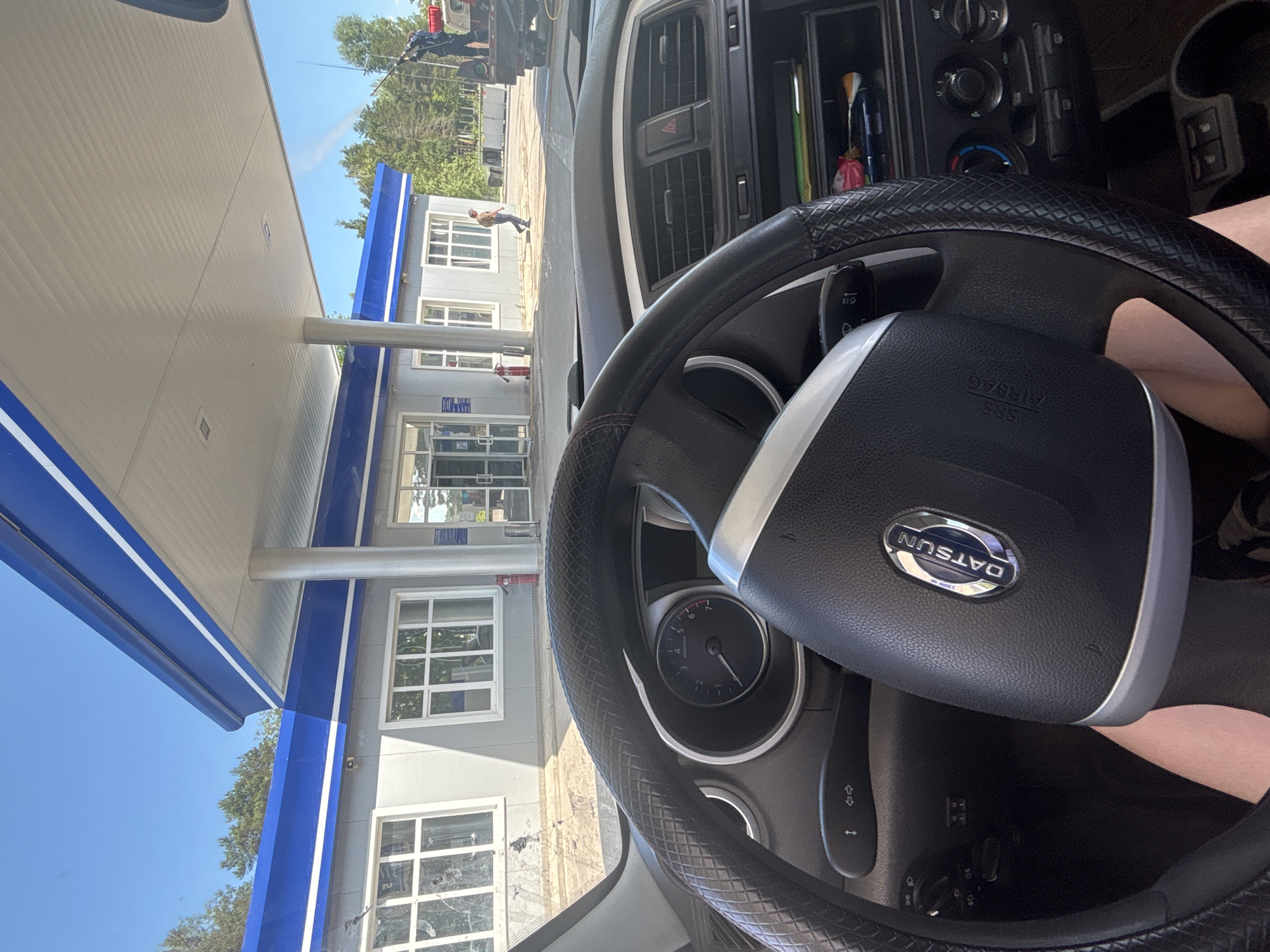 The image shows the interior of a car, looking out towards a gas station in Chaikovsky, Russia. The car's dashboard and steering wheel are visible in the foreground, with the DATSUN logo clearly displayed on the steering wheel. Beyond the car, the gas station has a blue and white canopy supported by pillars. The building of the gas station is light gray with white-framed windows. A person is seen walking away from the building towards the right side of the image. To the right of the gas station, a truck with a person working on it is visible. The sky is clear and blue, and trees are present in the background. The overall scene depicts a bright, sunny day at a gas station.