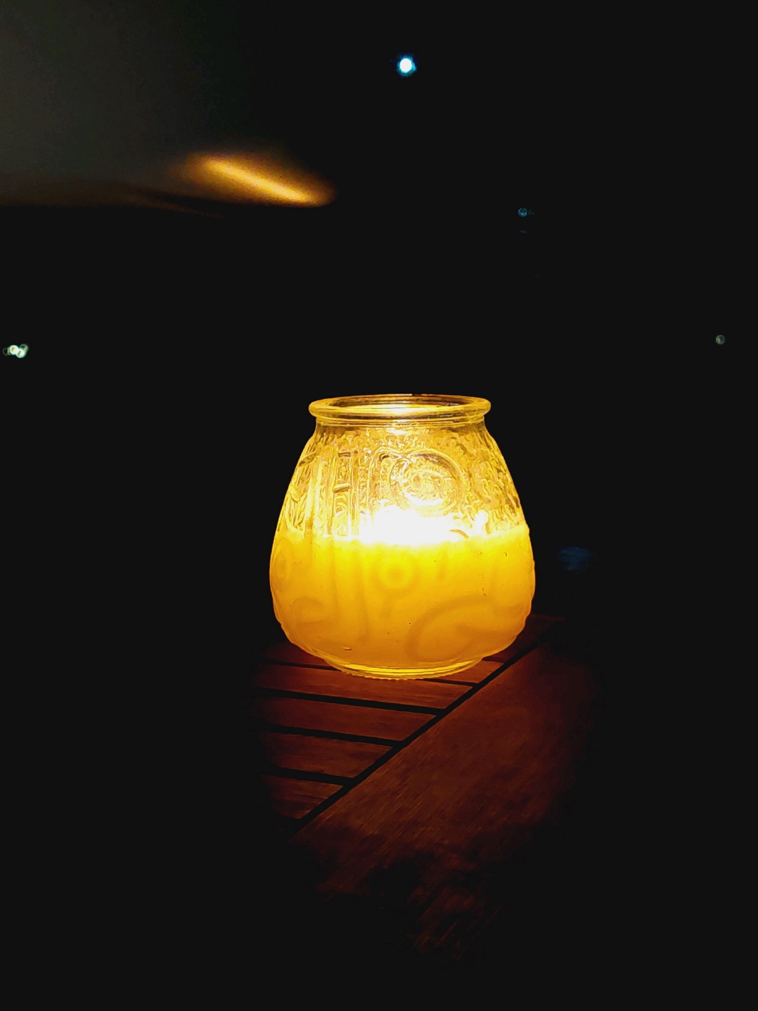 The image displays a close-up of a lit candle in a glass holder, placed on a wooden surface. The candle's flame emits a warm yellow light that illuminates the textured glass, revealing ornate patterns. The background is dark and out of focus, with a few distant lights visible, suggesting an outdoor setting at night. There are no people or visible interactions in the scene. The overall ambiance is serene and atmospheric. The location in Sozopol, Bulgaria, is not discernible from the visual information provided.