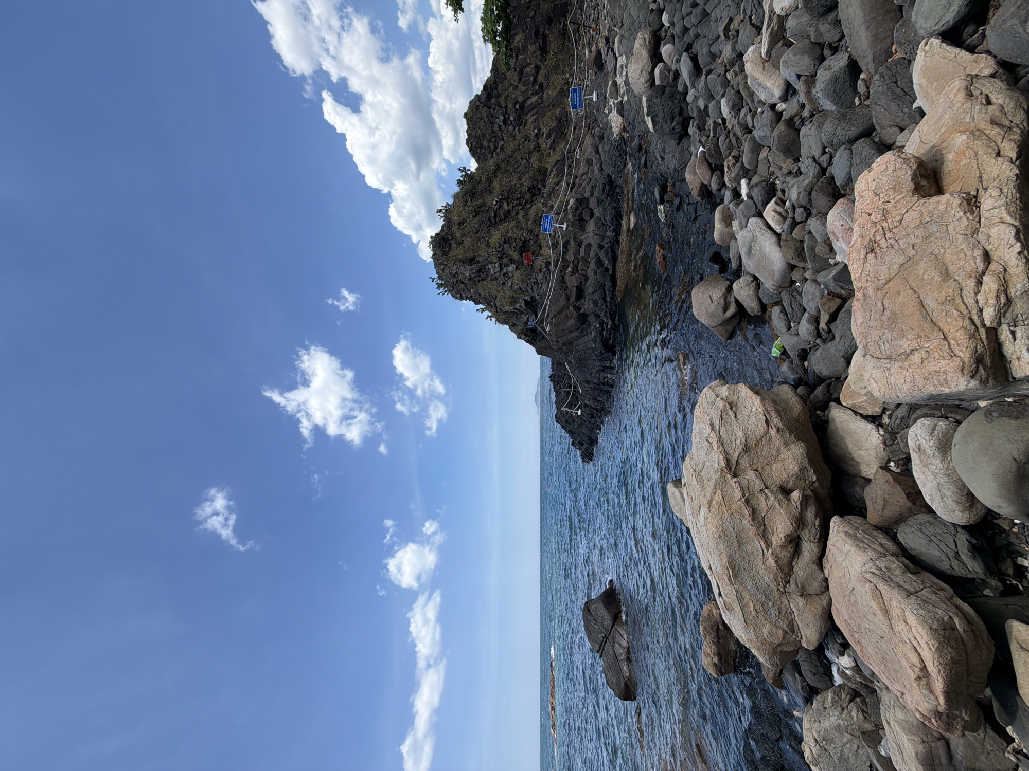 The image shows a rocky coastline with the sea and a bright blue sky. In the foreground, there are many large and small rocks of various colors, including tan, gray, and dark gray. Some of these rocks are smooth and rounded, while others are rough and jagged. The sea is a deep blue color, with gentle waves lapping at the shore. In the middle ground, there is a large, dark rock formation that juts out into the sea. This formation is covered in vegetation and has a path leading up to it, marked by a rope fence and several blue signs. In the background, the sea stretches to the horizon, meeting the sky under a partly cloudy sky with scattered white clouds. The overall scene is a natural landscape with a sense of rugged beauty. This location is likely a coastal area in Phú Lương, Vietnam, possibly a scenic spot or a nature reserve.