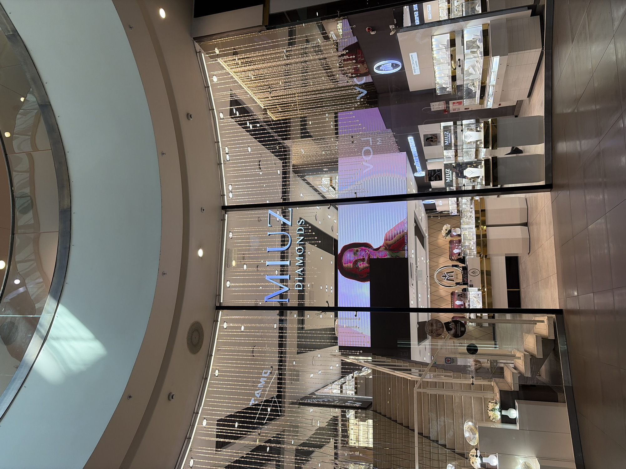 The image shows the interior of a shopping mall, likely in Saint Petersburg, Russia, focusing on a jewelry store called "MIUZ DIAMONDS." The store has a large, illuminated display window featuring a digital screen with a portrait of a woman. Inside, glass display cases exhibit jewelry, and mannequins are dressed to showcase necklaces and pendants. A staircase with a glass railing is visible to the left, leading to another level of the mall. The store's facade is adorned with hanging decorative lights. The lighting appears to be a mix of overhead mall lights and the store's internal illumination, creating a bright and upscale atmosphere. The overall scene is clean and modern, typical of a high-end retail environment.