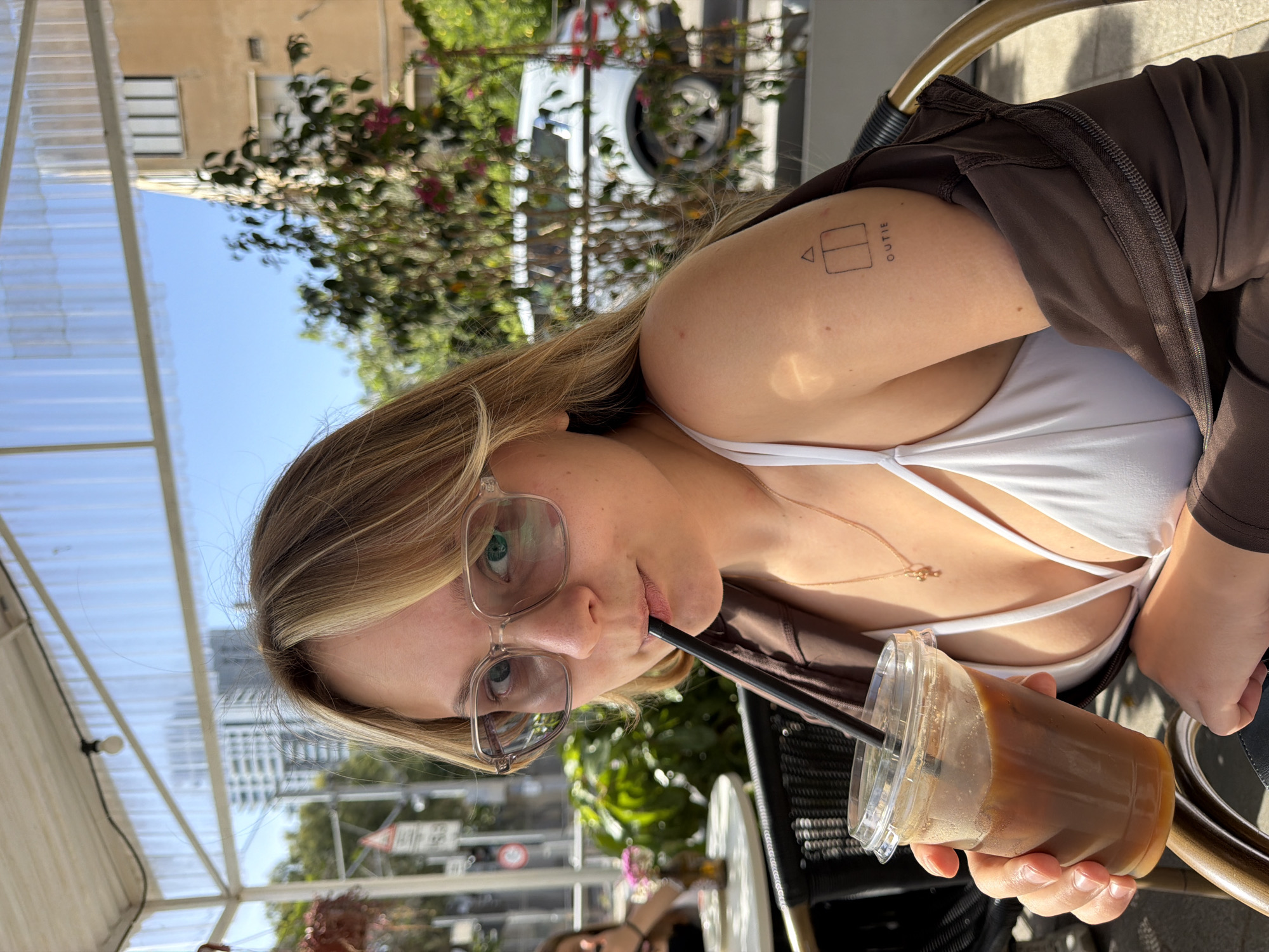 A young woman is seated outdoors, possibly at a cafe, drinking a beverage through a straw. She wears glasses, a white top, and a brown jacket. She has a tattoo on her upper arm. The background includes trees, buildings, and a covered outdoor seating area. The location appears to be in Bat Yam, Israel, based on the style of buildings, vegetation, and street signs visible in the background. The woman is holding a clear plastic cup containing an iced beverage.