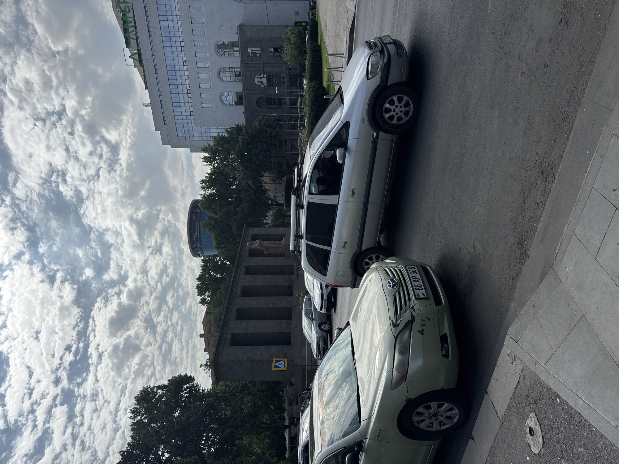 The image captures a street scene in Yerevan, Armenia, likely in the vicinity of the Cascade complex, given the architectural style of the building with arched windows in the background and the prominent monument with columns and a statue. The foreground features a light green Toyota Camry with the license plate "88 AP 868". To its right, a silver minivan with a roof rack is driving on the road. Several other cars are visible in the middle ground, and a pedestrian crossing sign is mounted on a pole. The environment is urban, with trees lining the street and a large modern building with the "ALAMERIA" logo visible in the distance. The sky is partly cloudy.