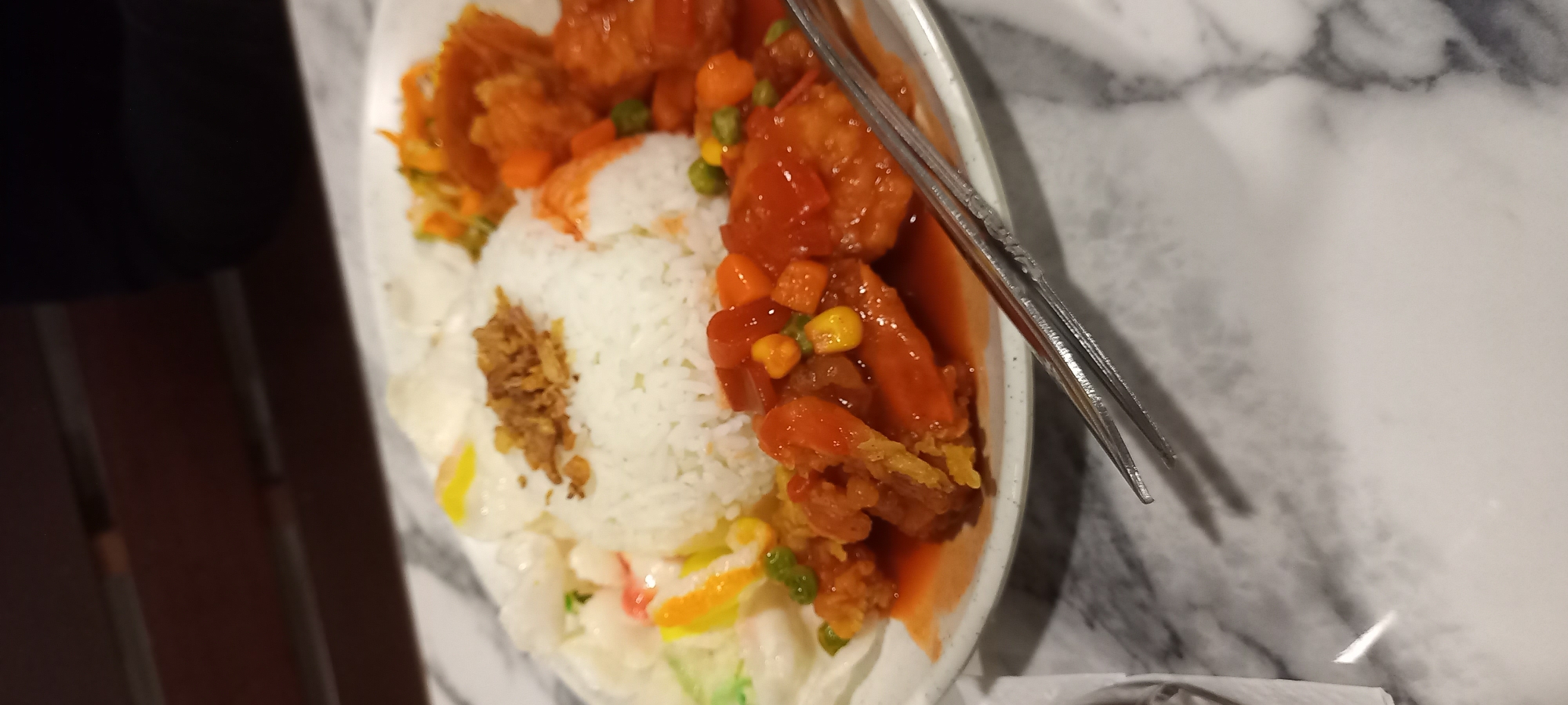 A plate of food is on a marble-patterned table. The plate contains white rice topped with fried onions, chicken pieces covered in a red sauce with diced carrots and corn, a salad with shredded carrots and lettuce, and what appears to be hard-boiled eggs. There are also some white crackers on the side. A pair of metal tongs rests on the table, partially over the plate. The background shows a dark wooden surface and a hint of a person's dark clothing. The food is typical of a meal served in a restaurant or home setting in Timbau, Indonesia.