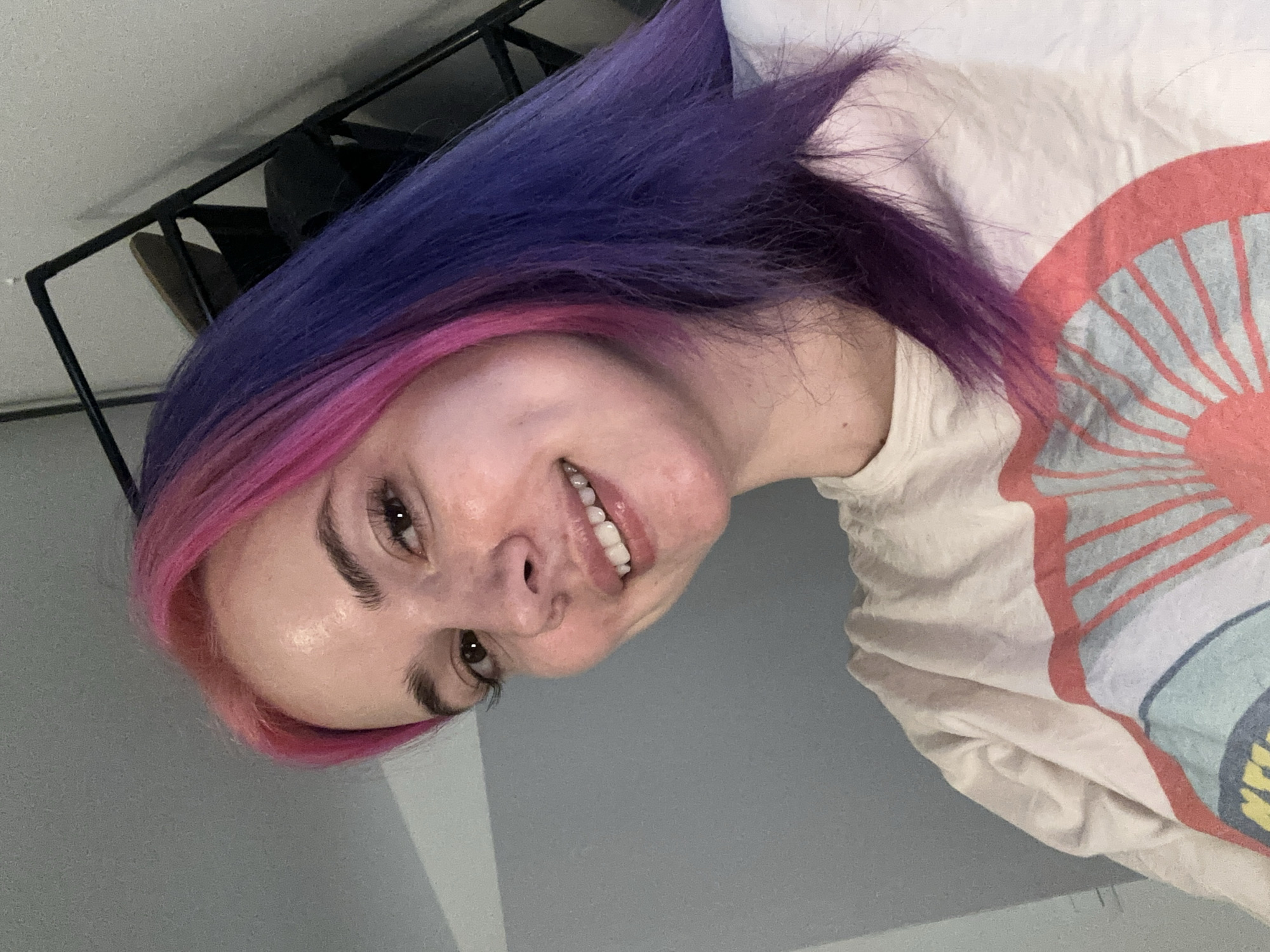 A young woman with dyed pink and purple hair smiles at the camera. She is wearing a white t-shirt with a colorful design. A black metal frame is visible in the background. The image appears to have been taken indoors. The location within City of Zagreb, Croatia cannot be determined from the image.