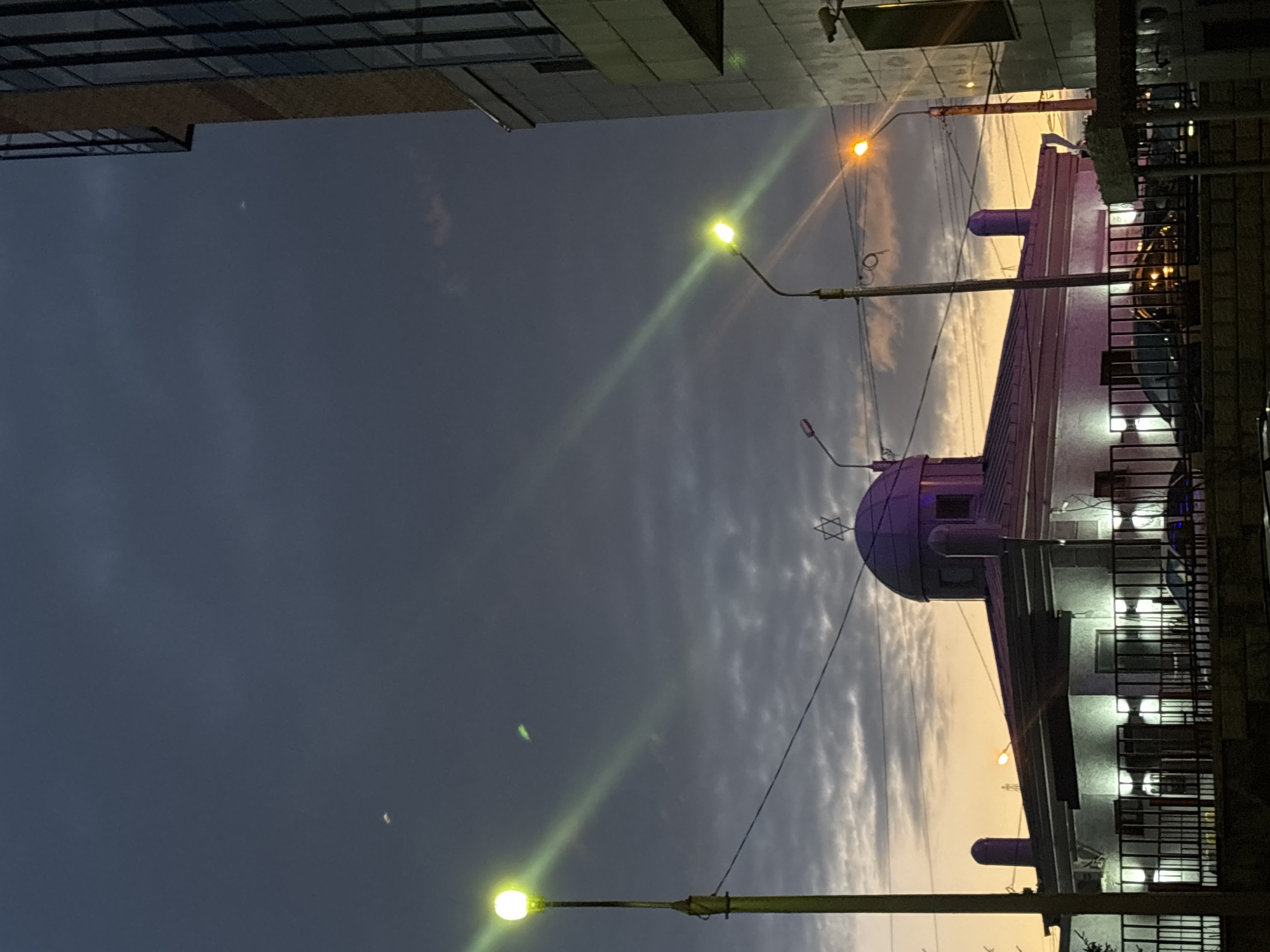 Here is a detailed description of the image:
**Setting:** An urban environment in Krasnoyarsk, Russia, likely at dusk or twilight. The photograph captures a view looking upwards, with buildings partially visible on either side framing a view of the sky.
**Objects:**
- A prominent synagogue, featuring a dome adorned with the Star of David, is positioned on the right side of the frame. The building is lit with internal lights shining through windows.
- Streetlights illuminate the scene with a yellow-green glow. Power lines are also visible against the sky.
- The sky displays a transition from dark blue at the upper left to a lighter, sunset-like gradient in the center. Streaks of clouds are visible within the sky.
**Architecture:** Modern high-rise structures are partially visible on the upper left side of the photograph.
**Overall Impression:** The photograph has an atmospheric quality, accentuated by the contrast between the building's lights and the fading daylight. The presence of the synagogue indicates the cultural or religious focus of the location.