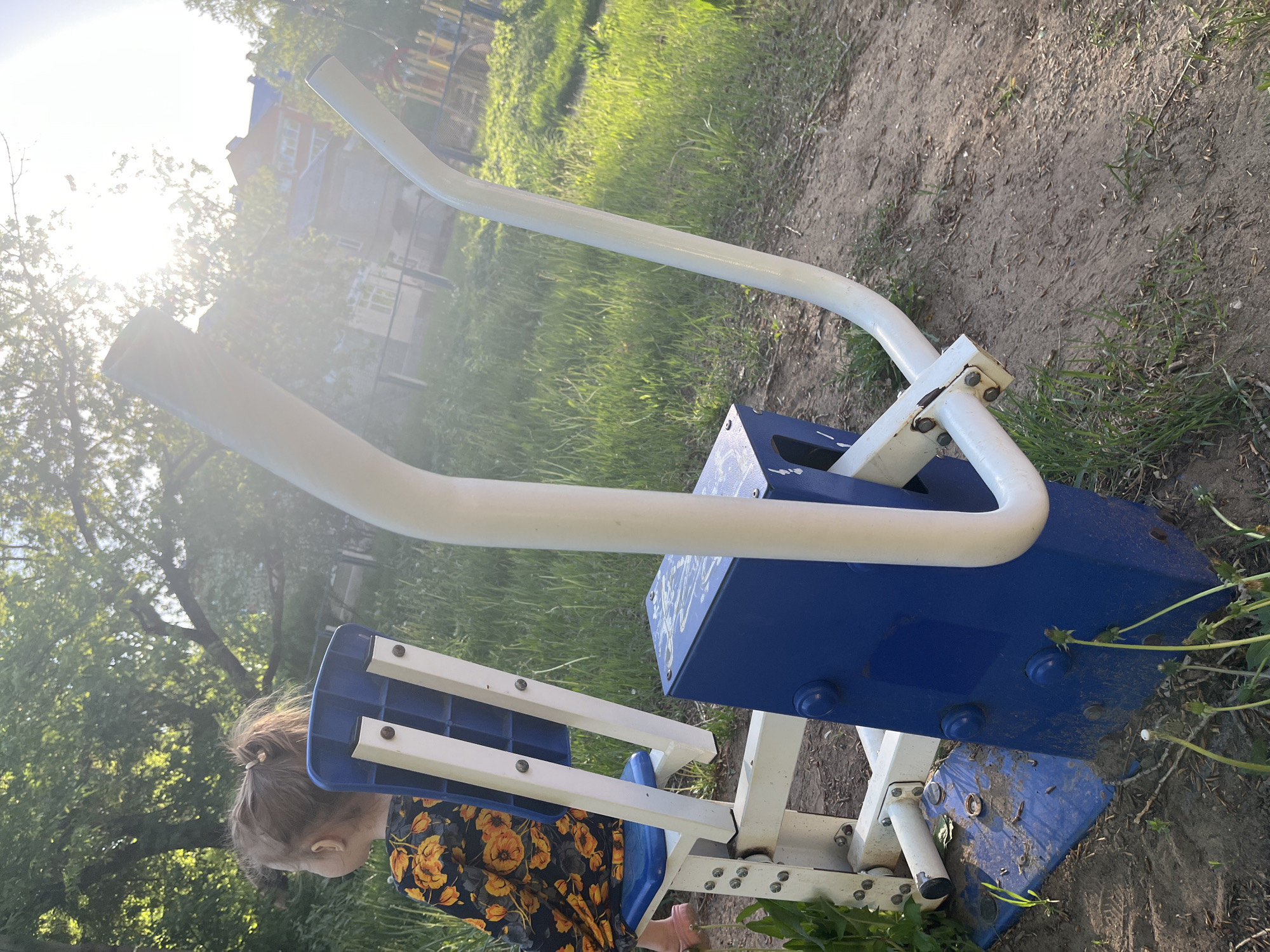 A slightly high-angle, close-up shot shows a piece of playground equipment, possibly an elliptical trainer, oriented horizontally, with a child positioned to the left. The equipment features a blue base with white handles and footrests. The paint on the base shows signs of wear.
The child, seen from the back, is wearing a floral-patterned shirt or dress. The child is not actively using the equipment.
The environment appears to be a grassy area, possibly a park or playground, with trees and a building visible in the background. The ground around the equipment is a mix of dirt and grass. Bright sunlight is present.