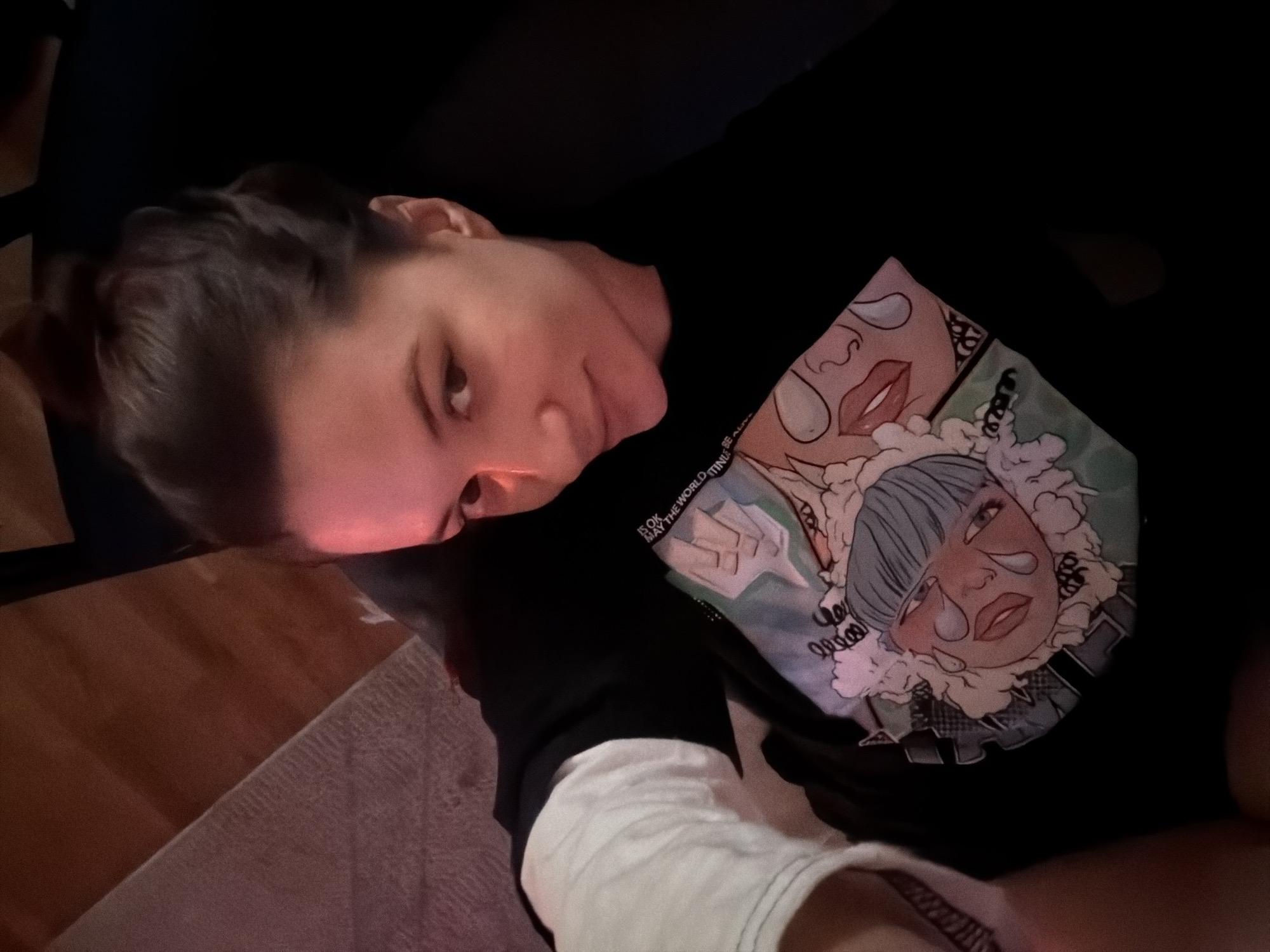 An individual is captured in an indoor setting, reclining or lying down, looking towards the viewer. The person has light brown hair pulled back, and their face is illuminated by a distinct red or pinkish glow, likely originating from a screen, suggesting a low-light environment such as evening or night.
They are wearing a black long-sleeved shirt featuring a colorful graphic design on the chest. The design depicts two stylized, crying faces with large tears, surrounded by white, cloud-like or fluffy elements. One face, prominently featured, has blue hair and a melancholic expression. Partially visible text on the shirt reads: "IS OK / MAY THE WORLD THIN". A white fabric, possibly a sleeve or undershirt, is visible on their arm.
The background is mostly dark, with glimpses of a wooden floor and a light-colored rug or blanket, indicating a casual home environment. The person's expression is neutral, contrasting with the emotional intensity of the shirt's graphic. While the provided context indicates Belgrade, Serbia, no specific visual cues in the image itself directly confirm this location.