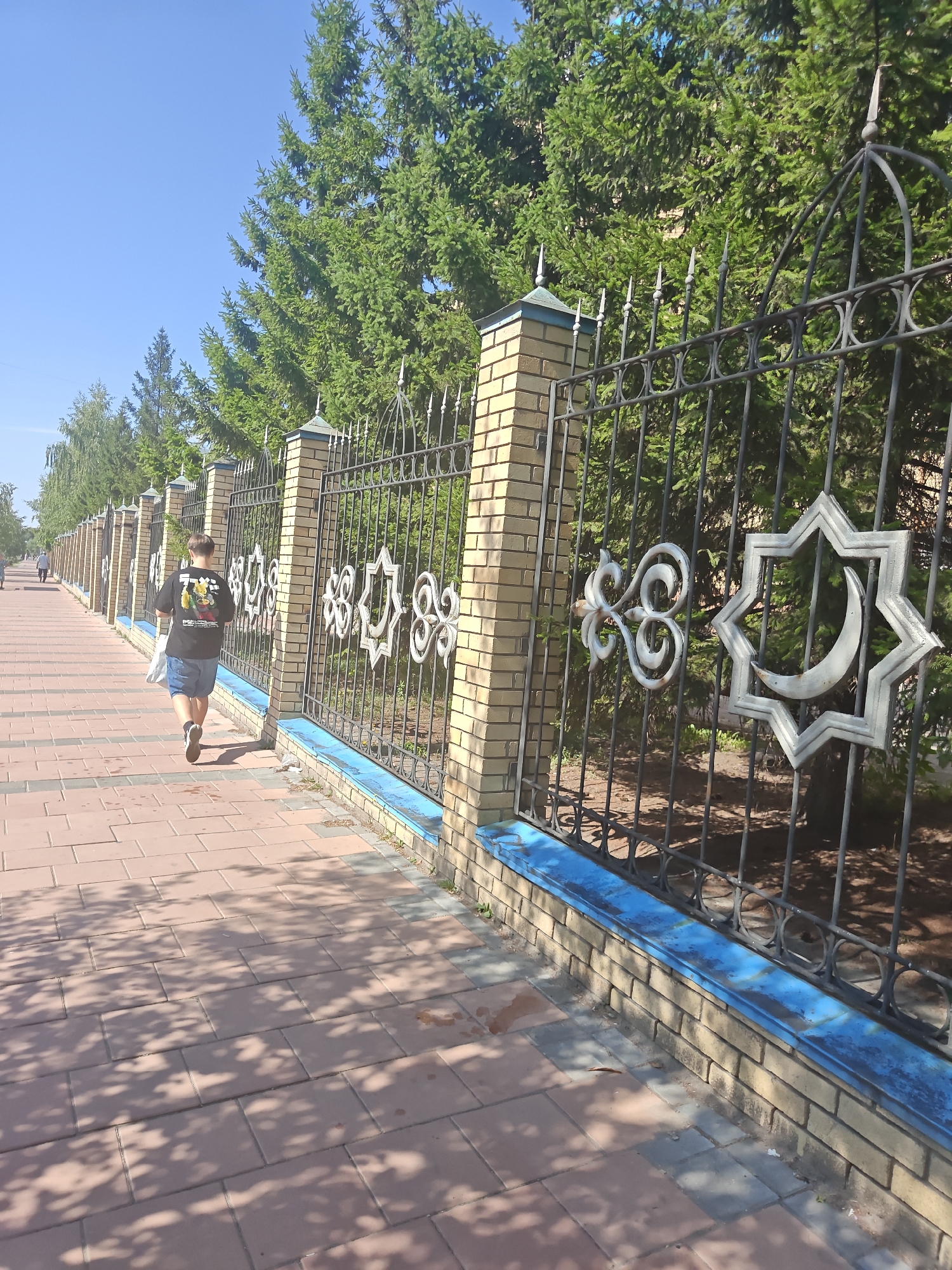 A young person walks away from the camera down a reddish-brown tiled pedestrian path. The person is wearing a black t-shirt with a graphic design and blue shorts, and carries a white bag. To the right of the path is a metal fence with ornate designs, including a star with a crescent moon. The fence is supported by brick pillars. Behind the fence, a row of green evergreen trees is visible, with a clear blue sky overhead. To the left of the path, the pedestrian walkway continues, with more trees and people in the distance. The scene appears to be a sunny day in an urban park or along a city street in Petropavl, Kazakhstan.