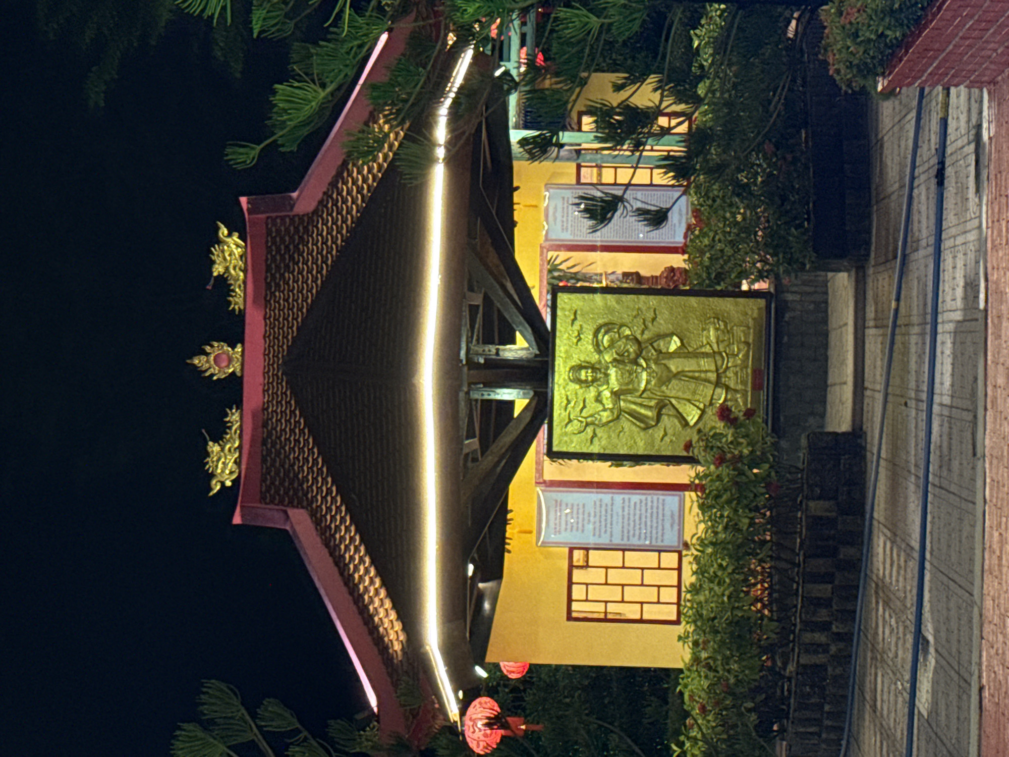 Captured in Bac Nha Trang Ward, Vietnam, the image presents an illuminated pagoda structure at night. The pagoda features a multi-tiered, brown-tiled roof with a red trim. Golden decorative elements adorn the roof edges. A bright strip of light runs along the center of the roof. Below the roof, a yellow wall with windows and inscribed plaques is visible. A square, golden bas-relief sculpture stands prominently in front of the pagoda. Dense greenery frames the structure on the right side. A brick walkway stretches towards the right side. The sky behind the pagoda is dark, providing a stark contrast to the brightly lit building.