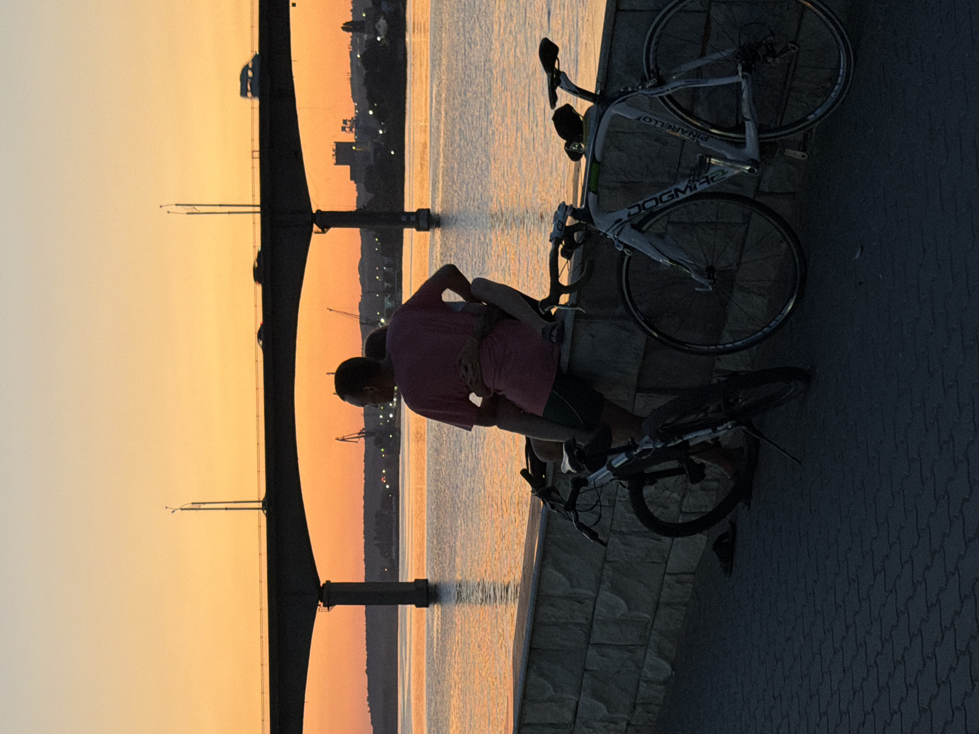 At sunset in Kamyshin, Russia, two figures are silhouetted against a vibrant orange sky as they embrace while sitting on a low stone wall overlooking a body of water. Two bicycles, one a dark mountain bike and the other a lighter road bike, are parked nearby on a paved walkway. In the background, a large bridge stretches across the water, with a few vehicles visible on its span. Further in the distance, industrial cranes and buildings line the shore, their forms softened by the fading light. The water reflects the warm hues of the sunset, creating a serene and romantic atmosphere.