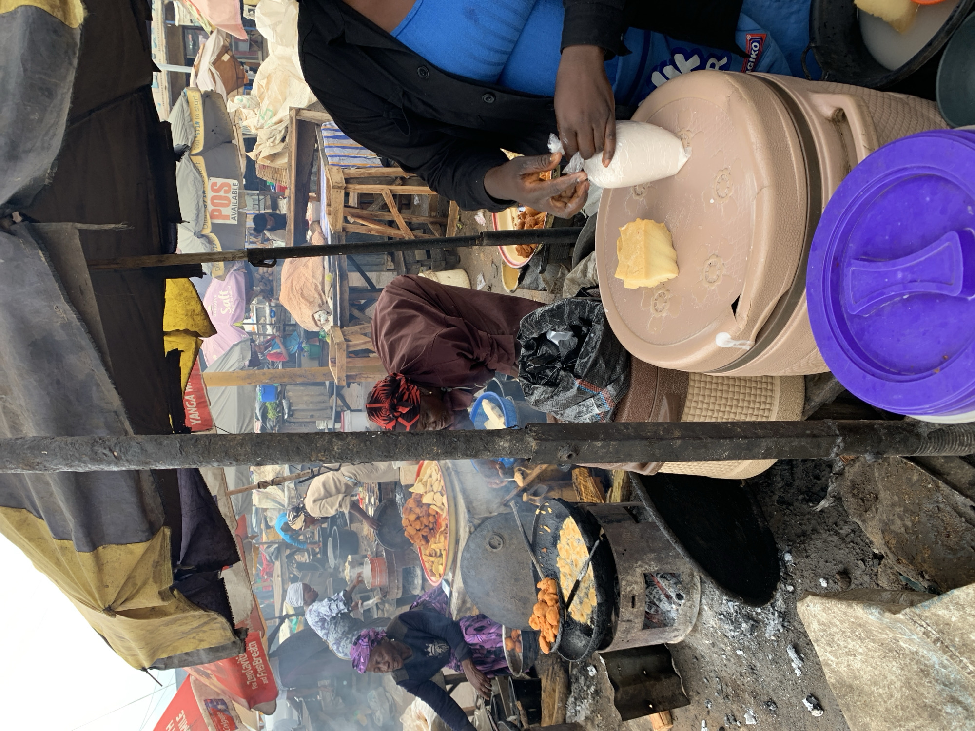 Here is a detailed description of the image:
**Overview:** The image depicts a bustling street food scene in Rafin Par, Nigeria. The perspective is from within a makeshift stall, looking out at other vendors and customers.
**People:** The image shows several individuals, including:
* A person preparing food in the foreground, wearing a blue shirt and black jacket. They are handling a white, dough-like substance.
* A woman bending over, appearing to be cooking near a pot on an open fire. She is wearing a maroon dress with a red-and-black head covering.
* Other vendors and customers are seen in the background, some cooking and some waiting.
**Subjects & Objects:** Key objects in the scene include:
* A large, beige thermos-like container with a purple lid, placed prominently in the foreground.
* A black pot on a charcoal fire, containing fried food. A metal skimmer is being used to remove the fried items.
* A metal stand with other fried foods on it.
* Several open fires for cooking
* Black and yellow canopies provide shade for the market stalls
* Various sacks and containers of ingredients are seen in the background.
* Advertising banners for brands like Colgate, Yanga W, and Dangote Salt
**Actions and Interactions:**
* Food preparation is the primary activity, with individuals cooking and packaging food.
* Transactions are likely occurring, with customers buying food from vendors.
**Environment:**
* The location is a busy market street in Rafin Par, Nigeria.
* The scene is crowded, with stalls and people packed closely together.
* Cooking is being done over open fires, creating smoke and heat.
* The structures are makeshift, suggesting a temporary or informal market setting.