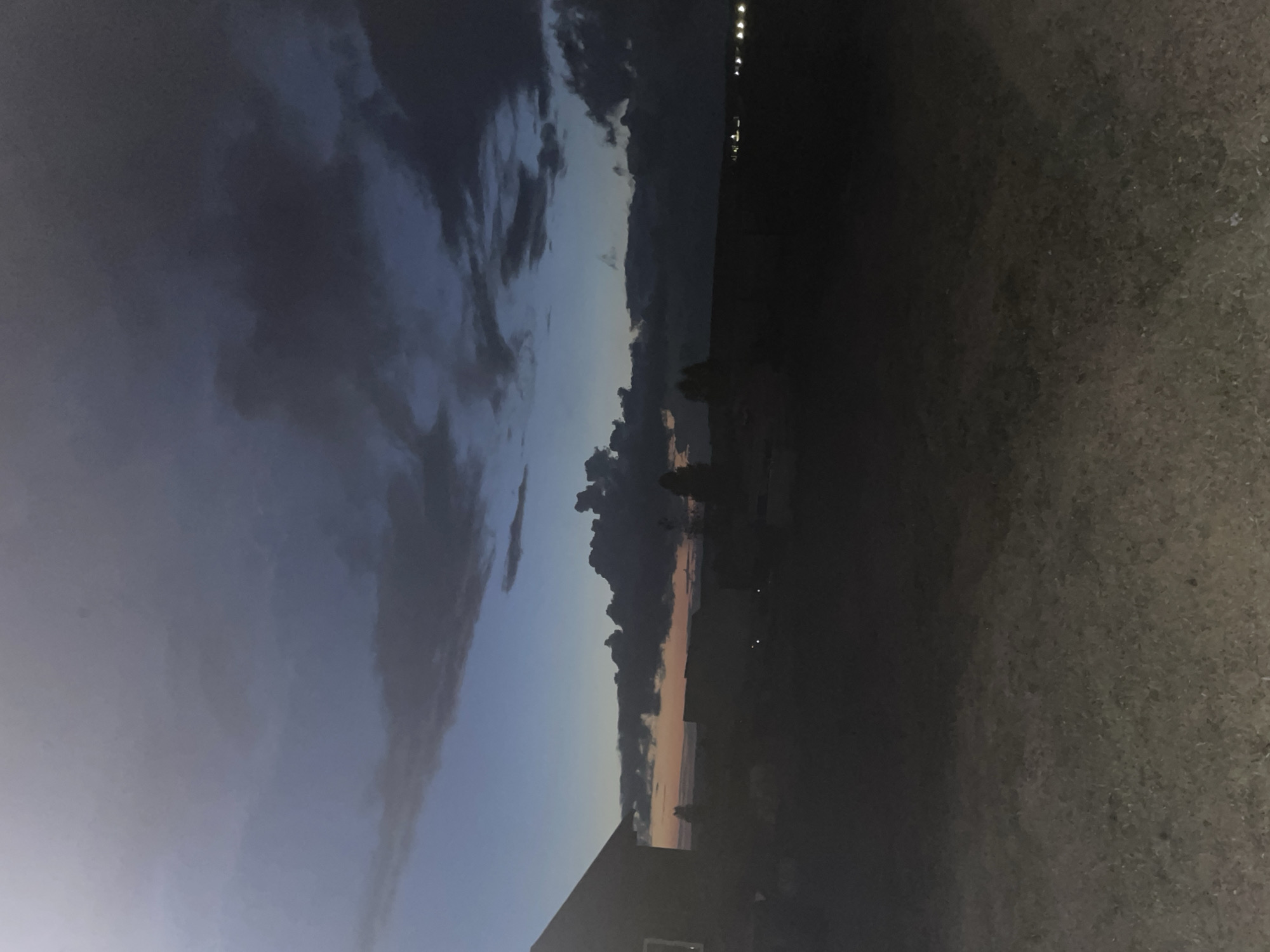 The image shows a twilight landscape in the city of Chulym, Russia. The sky is a deep blue with scattered dark clouds. A faint orange glow is visible on the horizon, suggesting either sunset or sunrise. In the foreground, there is a silhouette of a building, likely a house, on the left side of the frame. In the mid-ground, there are silhouetted structures that appear to be houses or other buildings, and some trees. A line of lights is visible in the far distance, indicating a populated area. The ground in the foreground is dark and appears to be gravel or dirt. There are no people visible in the image.