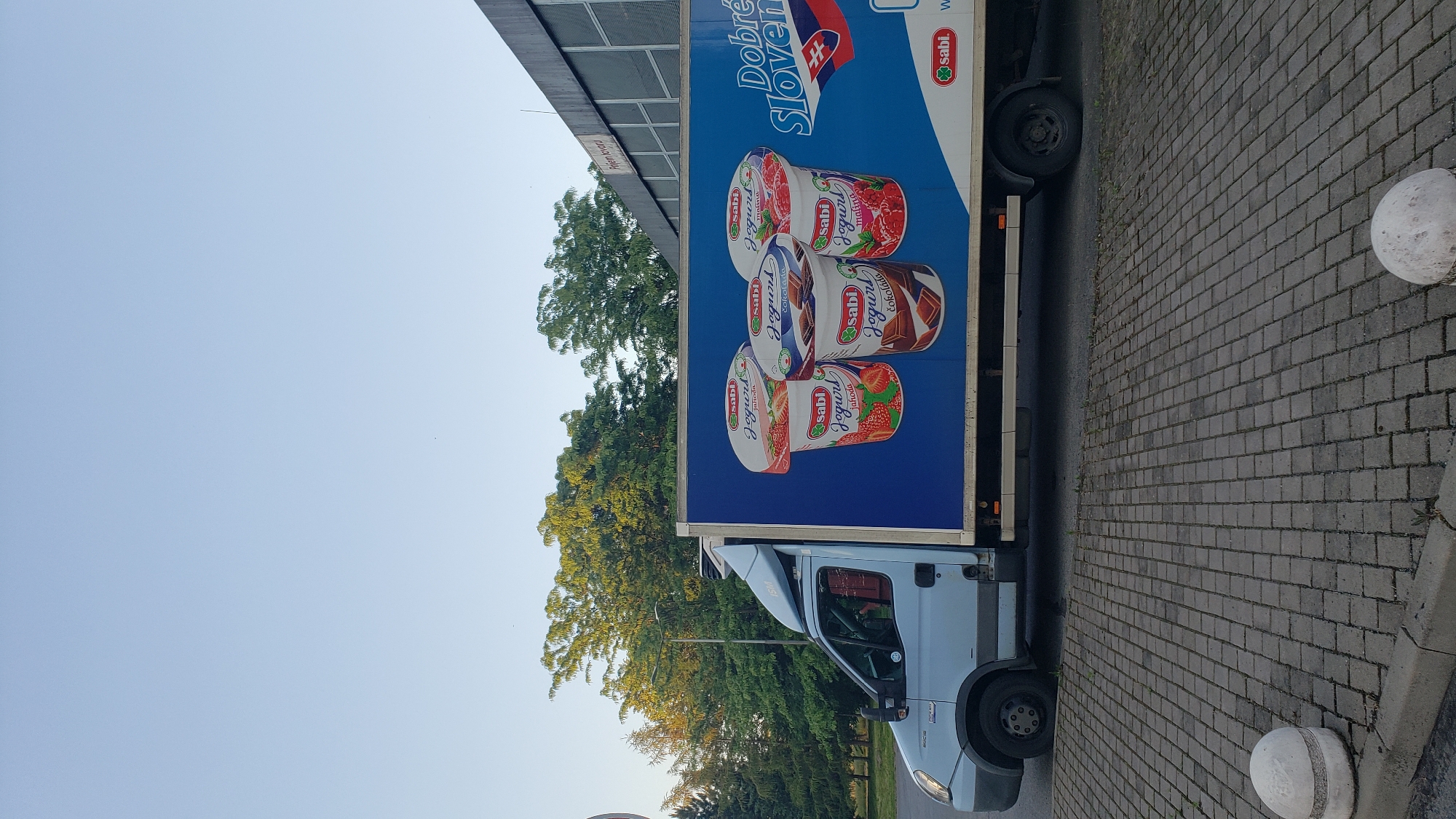 A light blue delivery truck is parked on a cobblestone surface. The truck's side features a large blue advertisement for "Sabi Jogurt." The advertisement displays three yogurt cups: one with strawberries, one with chocolate bars, and one with raspberries. The Slovak flag and the text "Dobré Slovensko" are also visible on the advertisement. The truck is parked next to a building with a modern facade. Trees with green foliage are visible in the background, suggesting an outdoor setting during the daytime, likely in a city in Slovakia given the flag and language on the advertisement.