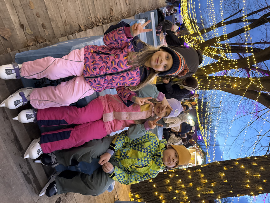 Three children are seated side-by-side on a light blue bench in an outdoor, evening setting. The child on the left, a girl with long blonde hair, wears a striped beanie with a pom-pom, a pink leopard-print winter jacket, light pink snow pants, and white ice skates. She smiles and displays a "peace" sign with both hands. The middle child, a girl, wears a light purple beanie, a pink, blue, and white color-blocked winter jacket, bright pink snow pants, and white ice skates. She also smiles, forming "peace" signs with both hands, one obscuring part of her face. The child on the right, a boy, wears a mustard yellow beanie, a green and blue geometric-patterned winter jacket, dark grey snow pants, and black ice skates. He looks slightly to his right with a neutral expression.
The children are dressed in winter clothing, suggesting cold weather conditions. The background features numerous bare trees illuminated by extensive strings of warm white fairy lights, which also crisscross overhead against a dark blue dusk sky. Other individuals, also in winter attire, are visible in the blurred background, some seated and others standing, alongside what appear to be market stalls or temporary structures. The ground in the immediate foreground consists of wooden planks with scattered dry leaves. The scene suggests a festive winter event, likely an ice-skating rink or winter market, located in the city of Unknown, Unknown.