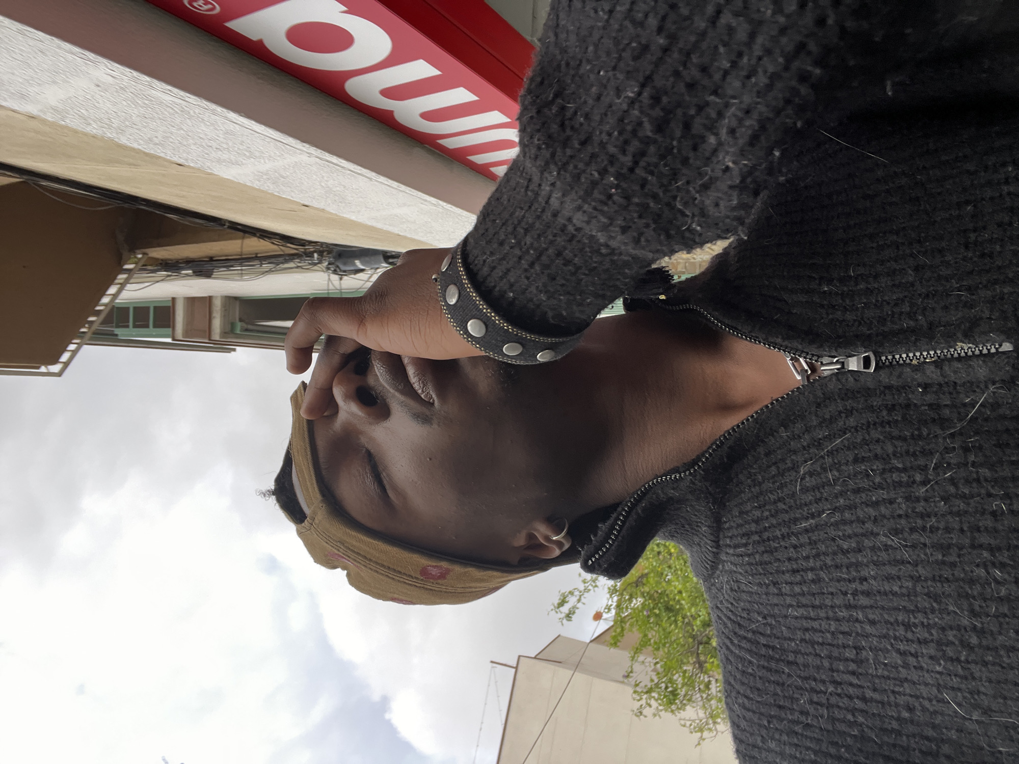 A man wearing a tan baseball cap, a black zippered sweater, and a studded bracelet is standing outside under a cloudy sky. His right hand is near his forehead. The bottom of a red and white sign is visible above him. Buildings and trees are in the background. The photograph appears to be taken at street level, with the camera pointing upwards towards the man. The location is likely Barcelona, Spain, considering the architectural style of the buildings.