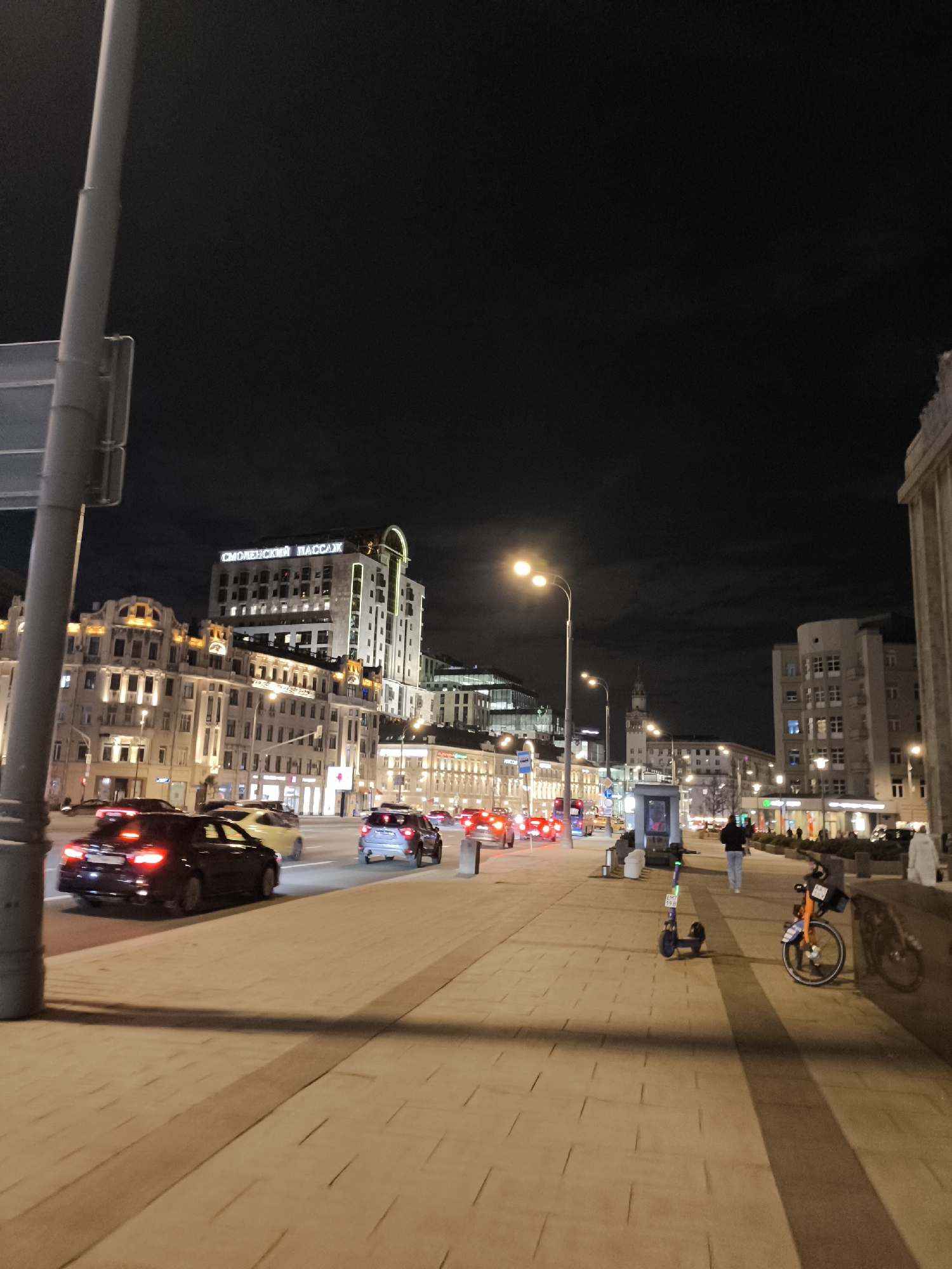The image captures a night scene on a bustling city street in Moscow, Russia. The sky is dark and overcast, indicating late evening or night.
The foreground on the left features a tall, light-colored street pole. To its right, a multi-lane road is visible with several cars. A black sedan with illuminated taillights is prominent in the immediate foreground, moving away from the camera. Other cars, including a yellow sedan and a gray SUV, are further down the street, their brake lights casting red glows.
On the right side of the image, a wide, paved sidewalk stretches into the distance. A few pedestrians are present; one person in a dark jacket walks away from the camera, and another in a light coat is further down. An electric scooter, purple and black, is parked on the sidewalk, along with an orange bicycle leaning against a low stone wall. Streetlights with bright yellow-orange glows illuminate the street and sidewalk.
Numerous multi-story buildings line both sides of the street. Many are well-lit, showcasing various architectural styles, from ornate, classic designs to more modern structures with glass facades. A prominent tall building in the mid-ground features illuminated text on its facade, clearly reading "СМОЛЕНСКИЙ ПАССАЖ" (Smolensky Passazh), confirming the location in Moscow. Another notable building with a distinctive spire is visible in the background on the right. The overall impression is one of a busy urban environment after dark, with continuous vehicular and pedestrian activity.