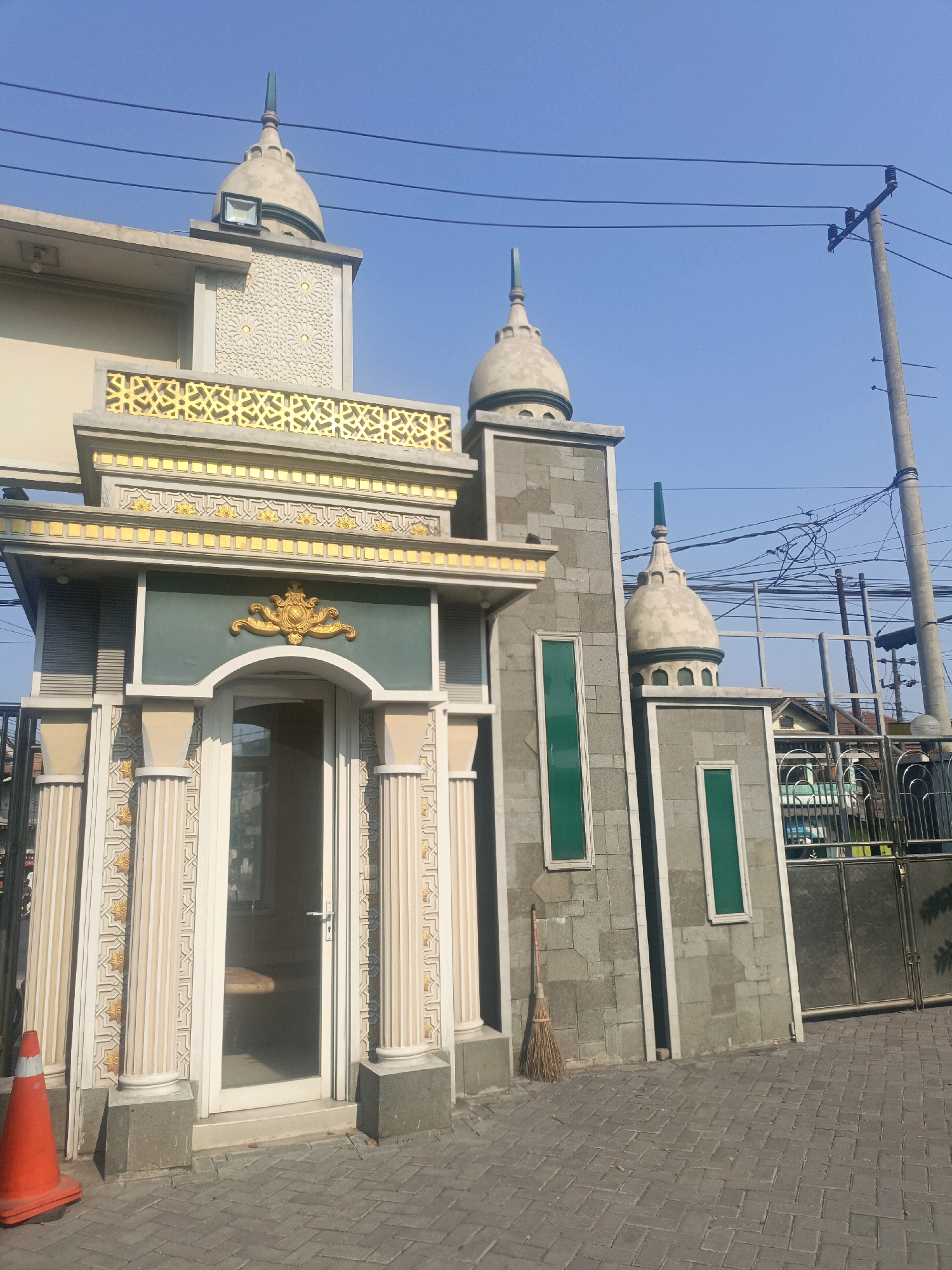 The image shows the facade of a building that appears to be a mosque or a religious institution in Gempol, Indonesia. The building features traditional Islamic architectural elements, including domes, minarets, and decorative patterns. The entrance is framed by columns and an arched doorway, with a glass door allowing a glimpse inside. To the right of the entrance, there's a grey brick wall with green rectangular windows and a broom leaning against it. A metal gate is visible to the far right, leading to an outdoor area. An orange traffic cone is placed near the entrance on the left. The scene is set against a clear blue sky, with power lines and a utility pole visible in the background, indicating an urban or semi-urban setting. The ground is paved with interlocking grey bricks.