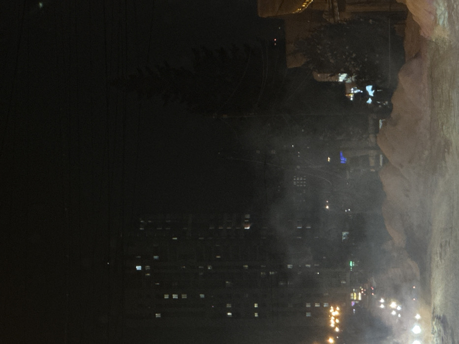A nighttime cityscape with numerous illuminated windows in tall buildings is partially obscured by a foreground feature that appears to be a rock or structure. Streetlights illuminate the base of the city, creating a hazy, glowing effect. Power lines are visible stretching across the dark sky.