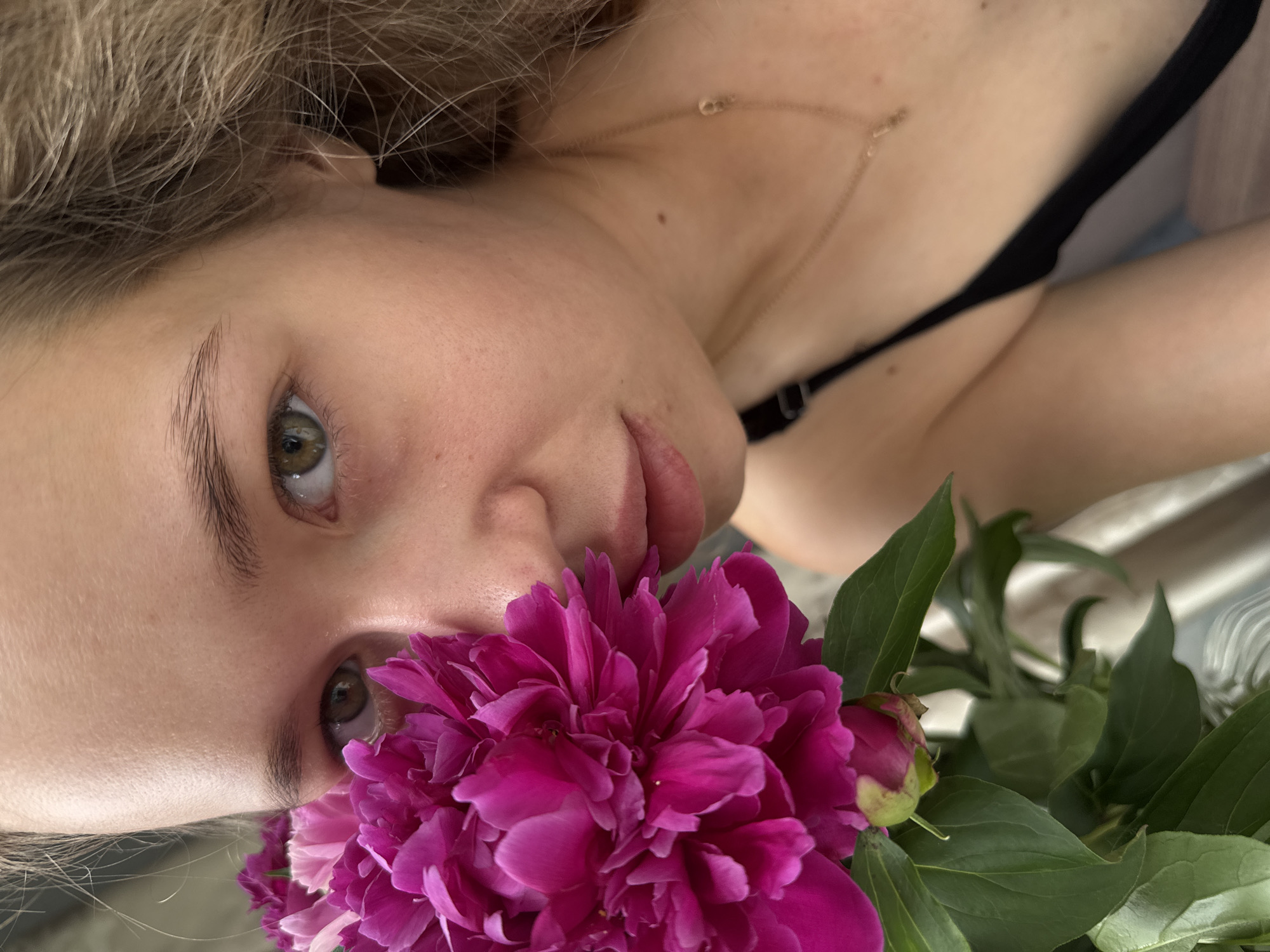 A close-up, waist-up portrait of a young woman with light brown hair and green eyes is shown. She is holding a large, vibrant pink peony flower near her face, obscuring part of her features. The woman is wearing a black camisole with a thin strap visible. A delicate gold necklace with a small pendant rests on her collarbone. Her skin appears fair and smooth, with some subtle texture visible. The background is softly blurred, with hints of light-colored fabric or curtains. The image is taken in a well-lit indoor environment, possibly a home, located in Chaikovsky, Russia.