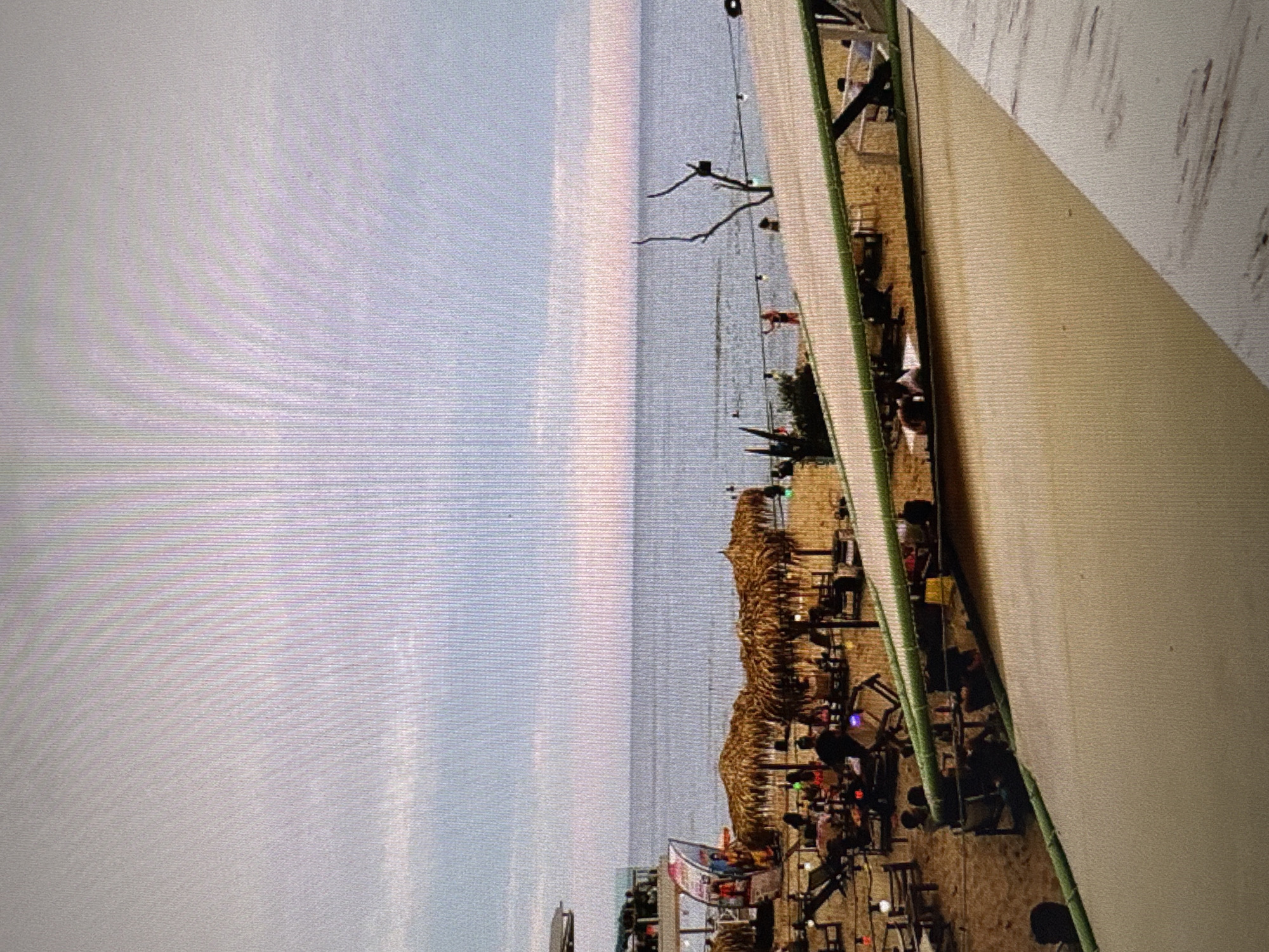 The image depicts a beach scene at what appears to be a seaside establishment in Varna, Bulgaria, likely during the late afternoon or early evening, given the lighting. The foreground is dominated by a light-colored canopy or awning extending into the frame, casting a shadow on the sandy beach below. Scattered across the sand are numerous people, some seated at tables under thatched-roof umbrellas, while others are standing or walking. Several lounge chairs are visible, indicating a recreational area.
The middle ground shows the sea, with a calm surface reflecting the soft hues of the sky. A few individuals are in the water. Along the shoreline, there are more seating arrangements and beach umbrellas, suggesting a vibrant, social atmosphere. A distinct skeletal tree branch stands near the water's edge, adding a natural element to the scene. In the background, the sky is a mix of pale blue and light pink, characteristic of a sunset or sunrise. The overall impression is that of a well-utilized beach resort or club.