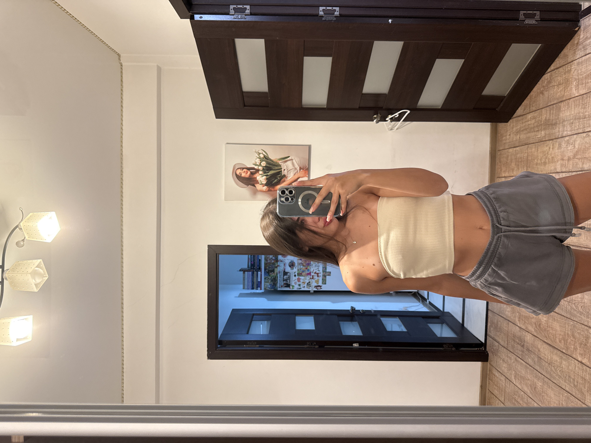 A young woman with long brown hair takes a mirror selfie in a dimly lit room. She is wearing a cream-colored strapless top and gray athletic shorts. Her phone, a silver iPhone with a prominent camera array, is held up to capture the reflection. Behind her, two dark wooden doors are visible, one slightly ajar revealing a glimpse of a refrigerator covered in various items. To the right of the open door, a framed picture of a woman wearing a hat and holding a bouquet of tulips hangs on the wall. The floor is made of light brown wooden planks. A string of decorative lights is mounted on the ceiling. The room appears to be residential.