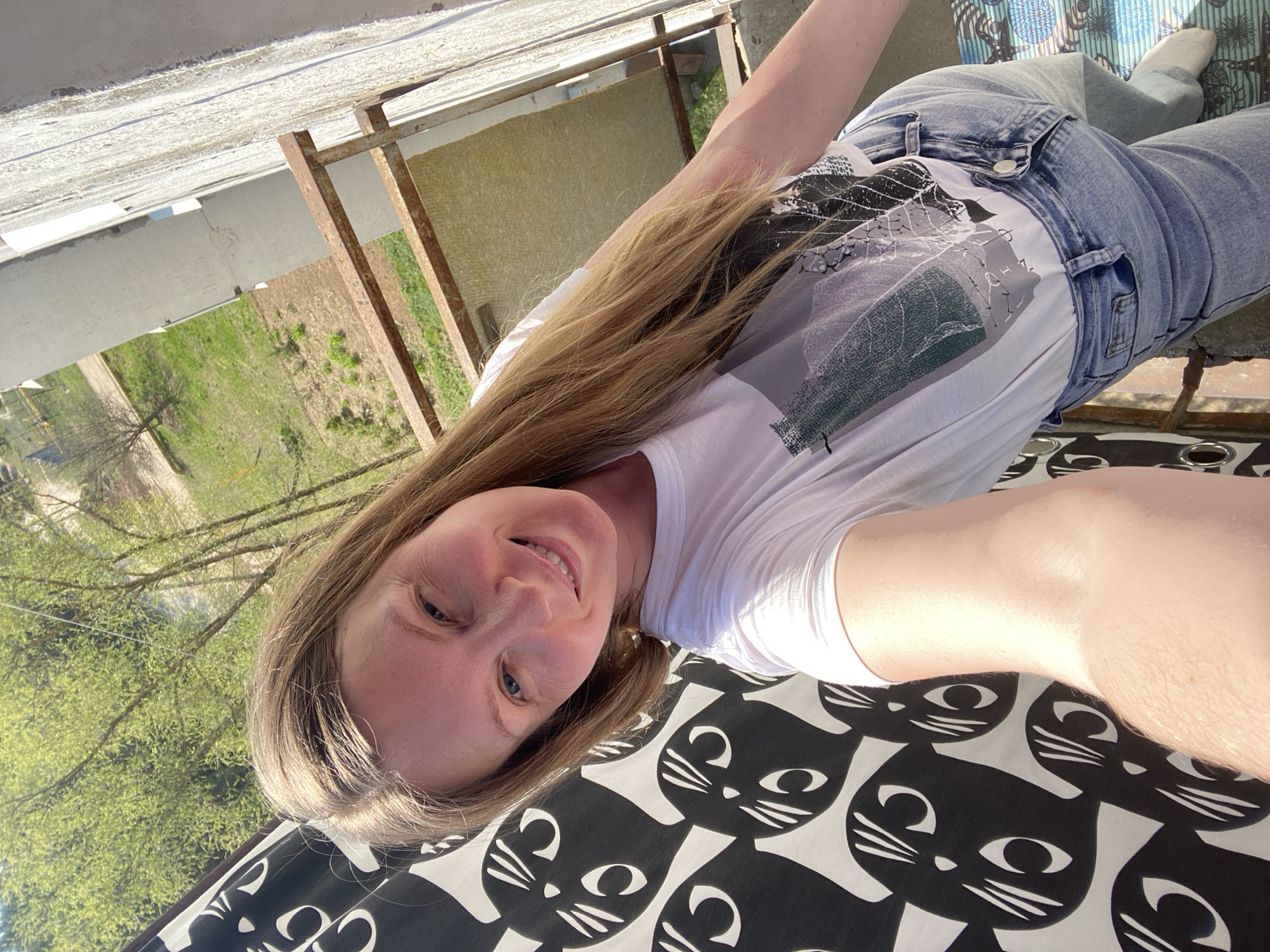 A woman is lying on a black and white patterned blanket with cat designs. She is wearing blue jeans and a white t-shirt with a leaf print. She has long, light brown hair and is smiling at the camera. The background shows a building with a metal structure next to a green field with trees. Based on the prompt, this location is in Noviy, Russia.