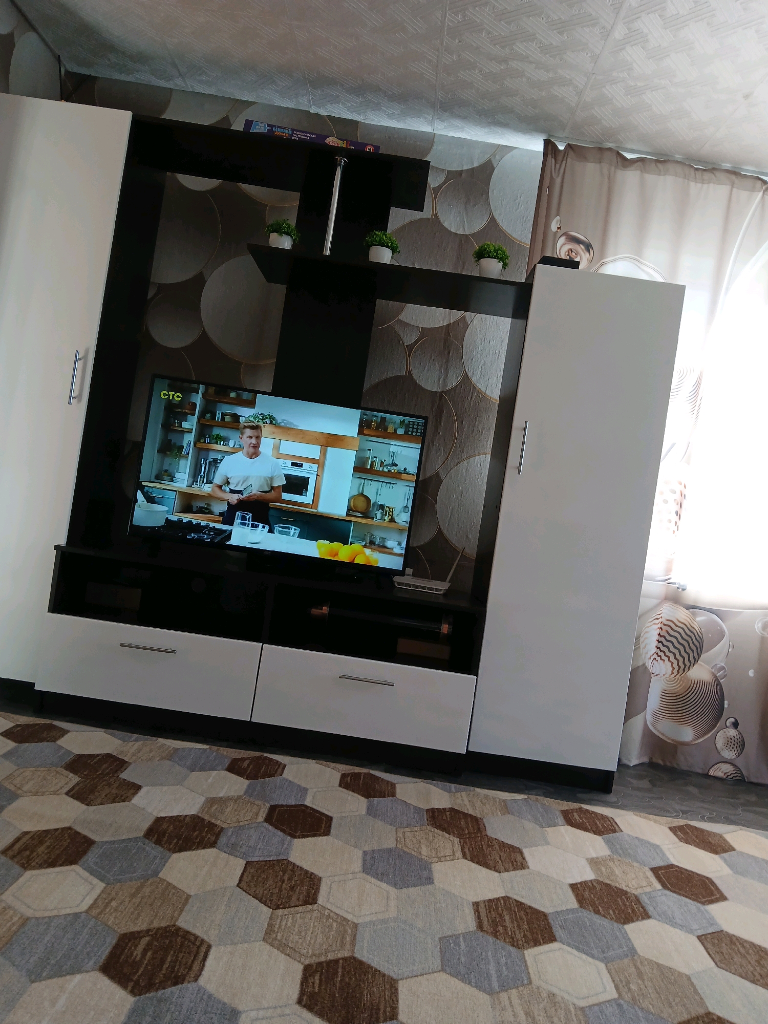 A wide shot captures a living room in a dwelling. The room is furnished with a large media console featuring a television screen displaying a cooking show. The console is flanked by two tall, white cabinets. The walls are decorated with a circular, textured wallpaper in shades of grey and beige. A light brown and beige hexagonal patterned rug covers a portion of the floor, which appears to be tiled. Curtains hang on the right side of the frame, partially obscuring a window. The overall impression is of a lived-in space.