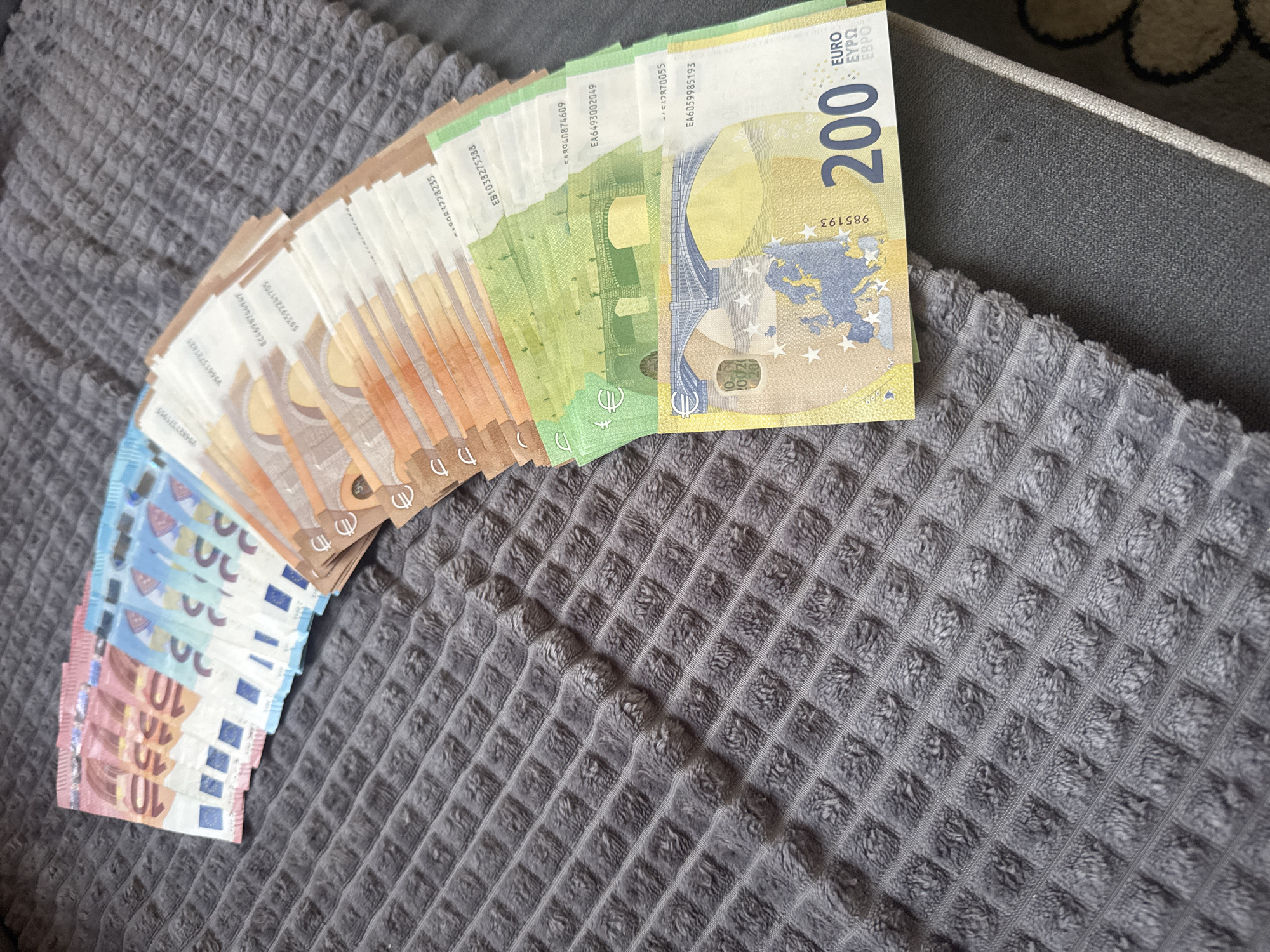 The image shows a fan-like arrangement of euro banknotes on a gray, textured fabric surface. The denominations of the banknotes visible are 10, 20, 50, 100, and 200 euros, arrayed in ascending order of value from left to right. The edges of the banknotes are staggered, revealing different sections of each bill. The image does not contain information about Vetren Dol, Bulgaria, or any people, subjects, actions, or interactions.