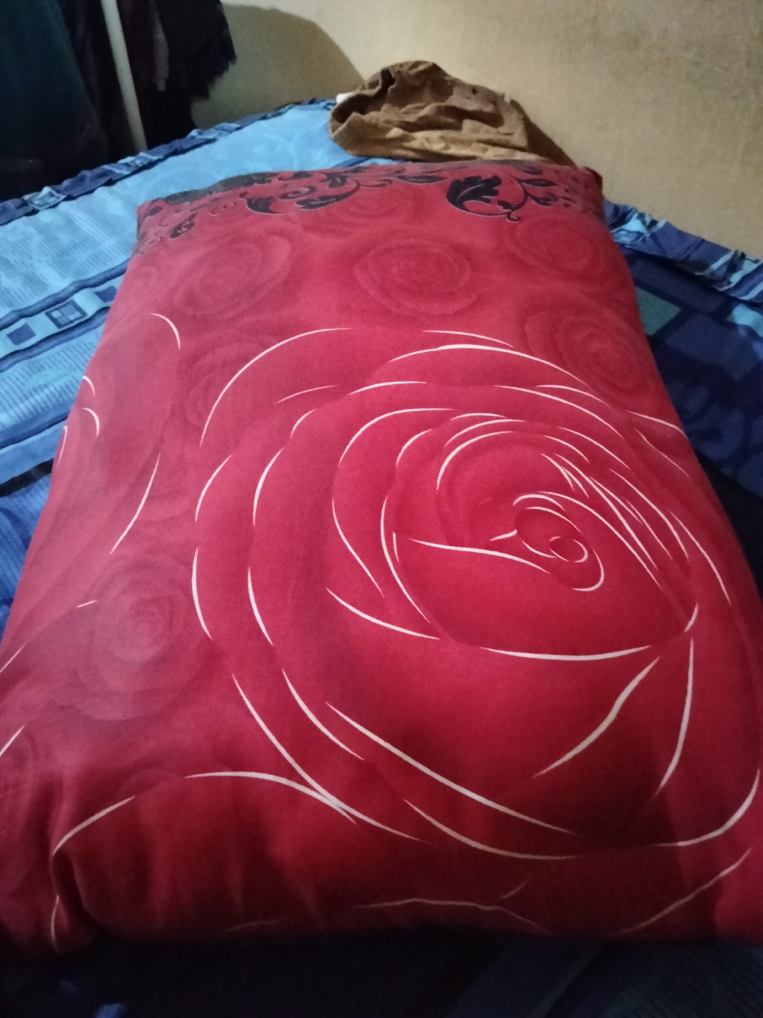 A red pillow with a white rose pattern is placed on a blue and white striped bed. The pillow is partially covered by a brown garment at the top. The scene appears to be indoors, likely a bedroom. The image is taken from a slightly elevated angle, looking down at the pillow.