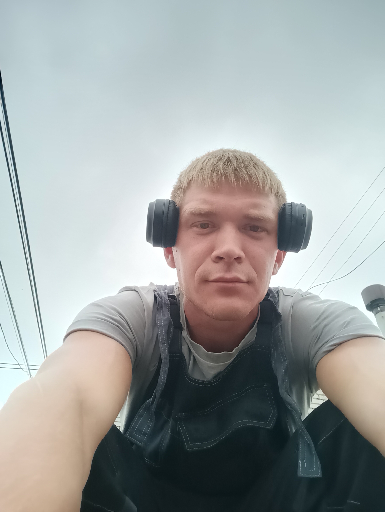 A young Caucasian male with short, light-colored hair and headphones over his ears is pictured from a low-angle, close-up perspective. He is wearing a gray t-shirt and dark overalls. The background consists of a pale, overcast sky and overhead wires, suggesting an outdoor urban setting. His arms are visible in the foreground, and he appears to be looking directly at the camera.