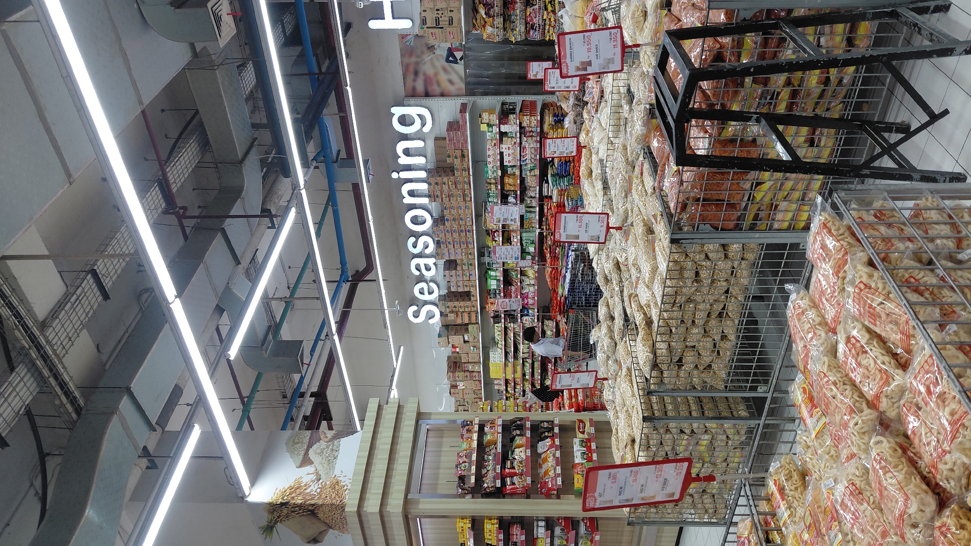 The image shows the interior of a grocery store in Purwokerto, Indonesia, specifically in the "Seasoning" aisle. The aisle is lined with shelves stocked with various food products, including bags of snacks displayed in metal cages in the foreground. In the middle ground, a man in a white shirt is seen pushing a shopping cart. The ceiling features fluorescent lighting and exposed ductwork. Signs indicating prices and product names are visible.
