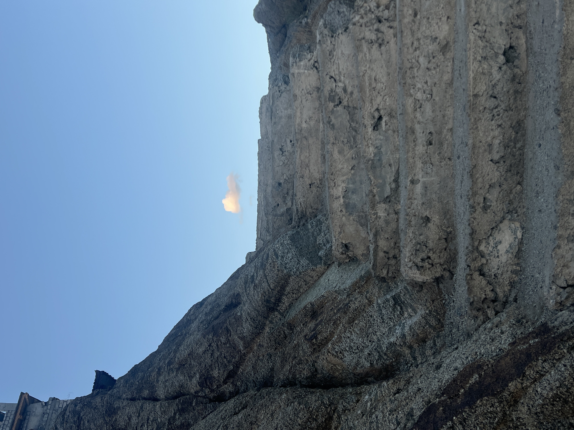 The image shows an outdoor scene with a steep, rocky incline to the left, partially covered by a rough, dark-colored material, possibly stone or a rocky outcrop. To the right of the incline, a series of rough, unfinished concrete stairs ascend upwards, appearing weathered and eroded. The sky is a clear, pale blue with a single, small, light-colored cloud positioned in the upper right quadrant. In the far upper left corner of the image, a small portion of a building with traditional architecture, featuring a dark, curved roofline and some lighter colored walls, is visible perched atop the rocky incline. This building's placement suggests it is situated at a high elevation. The overall environment appears to be arid and rugged, consistent with a mountainous region. This scene is located at the Leh Palace in Leh, India, known for its historical significance and commanding views of the surrounding landscape.