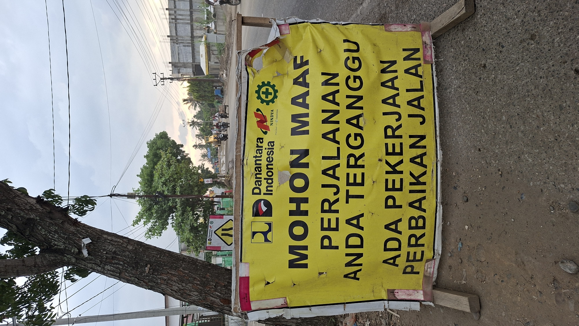 An outdoor, eye-level shot captures a yellow banner displaying a message in black lettering, positioned along a dirt and asphalt road. The banner, slightly torn and weathered, is supported by wooden posts. Above the banner, a large tree trunk with textured bark stands to the left. In the background, power lines crisscross the sky, and a construction site is visible with buildings, a yellow excavator, and several motorbikes with riders on the road, suggesting ongoing work in the city of Keudebayu, Indonesia. The sky is overcast, indicating a cloudy day.