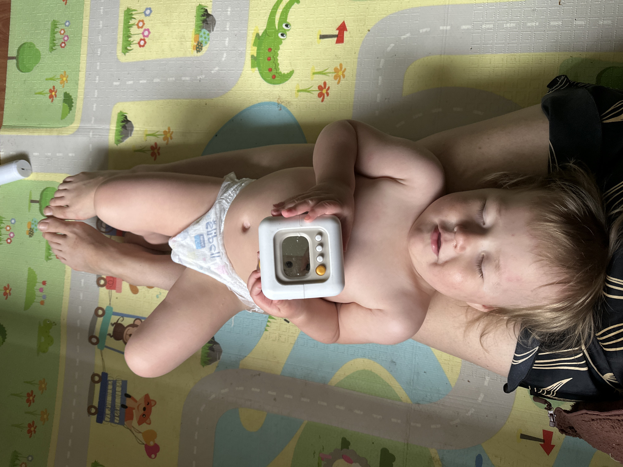 A baby, wearing a diaper, lies on a person's lap on a colorful play mat. The baby holds a white timer with a small digital display and several buttons. The play mat features a road pattern with various cartoon-like animals and plants, including a green alligator. The person's legs are visible, as is a portion of their clothing. A white cylinder is visible in the background. The location is not identifiable from the image.