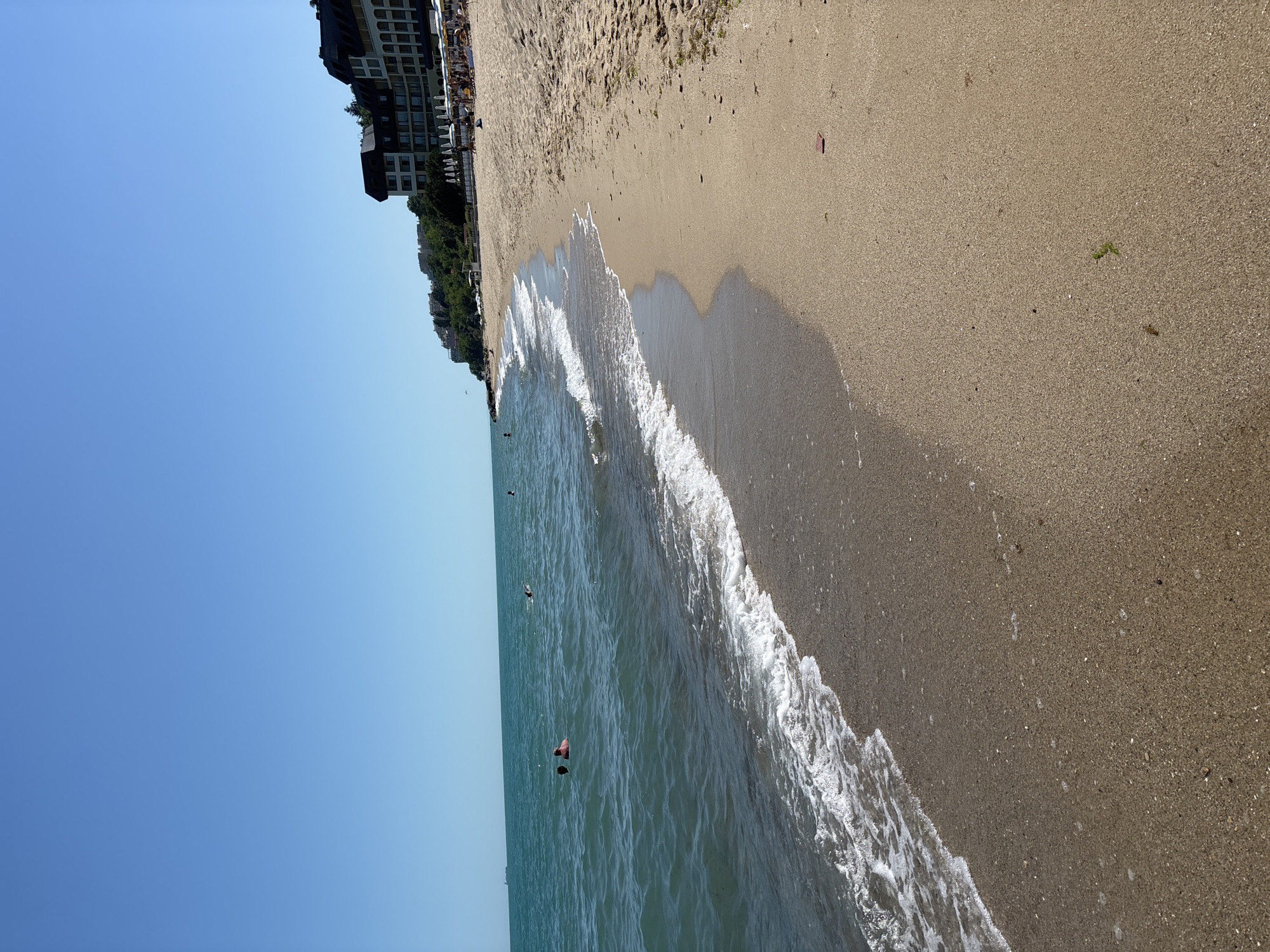 The image shows a sandy beach on a sunny day in Varna, Bulgaria, with the Black Sea on the left and a resort hotel on a hill in the background. The sea is calm with gentle waves lapping at the shore. Several people are visible swimming or wading in the water. On the beach, there are some beach umbrellas and loungers, with more people visible further up the beach near the hotel. The sky is clear and blue.