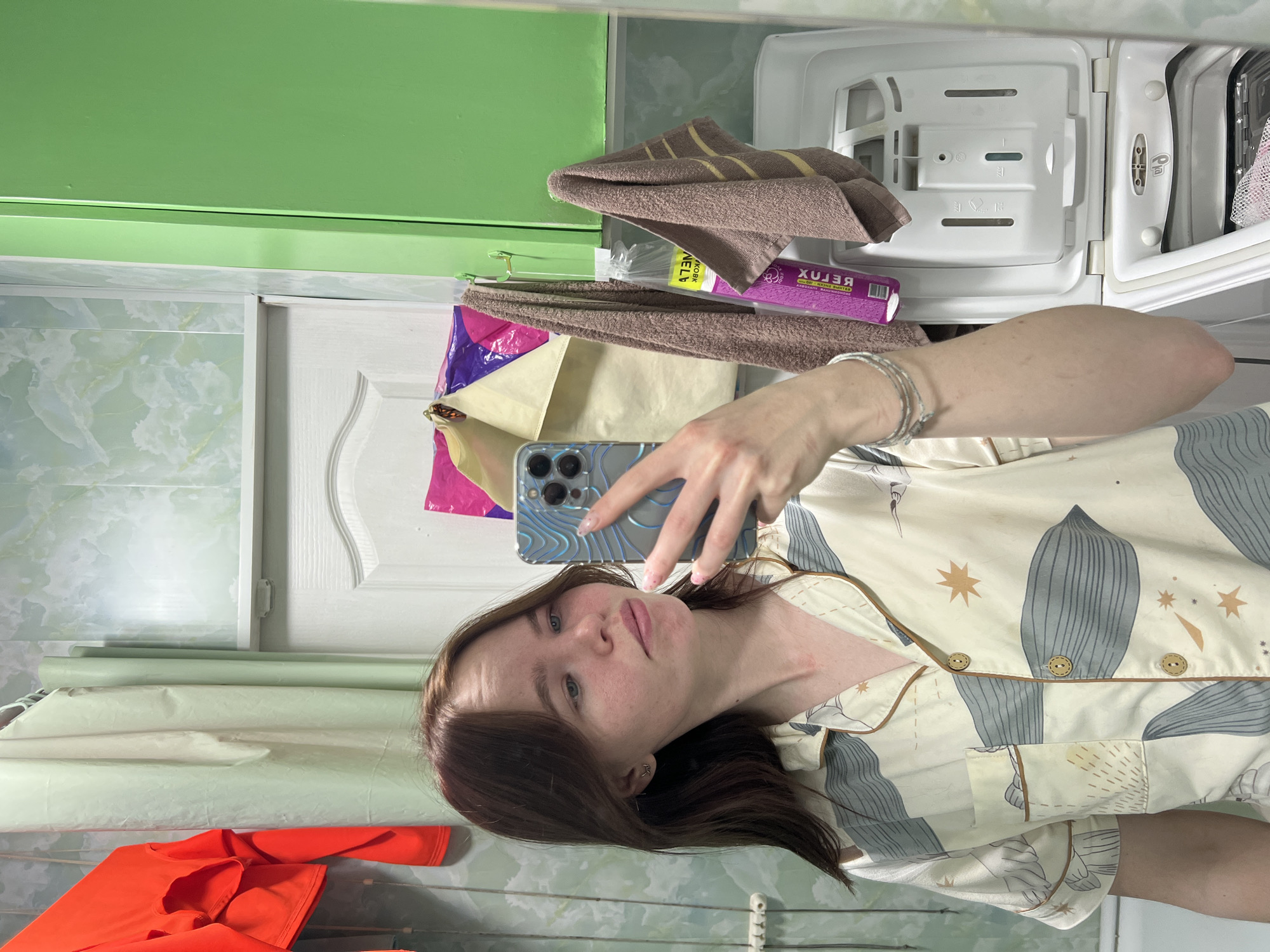 An eye-level mirror selfie shows a young woman with brown hair and fair skin. She is wearing a light-colored, long-sleeved top patterned with blue/grey leaf-like shapes and small orange stars, featuring brown piping and buttons. On her right wrist, she wears a silver bracelet. She holds a smartphone with a textured blue/silver case, showing light pink manicured nails. Her expression is neutral as she looks towards the camera.
The reflection in the mirror reveals an indoor setting, likely a laundry room or bathroom in Chaikovsky, Russia. To the left, a bright green painted surface is visible above a wall covered in light green and white marbled tiles. A white cabinet door with a curved panel is also seen. To the right, the open white plastic detergent drawer of a washing machine is prominent, displaying various compartments and symbols. Two folded brown towels with yellow stripes are placed near the washing machine drawer. Below them, a purple package with inverted text, including "ХБЛЭЯ" (which reads as "BELUX" when inverted) and "WKOBA," rests next to a beige paper bag. In the foreground, outside the reflection, a folded orange garment, possibly a t-shirt, is visible on a surface.