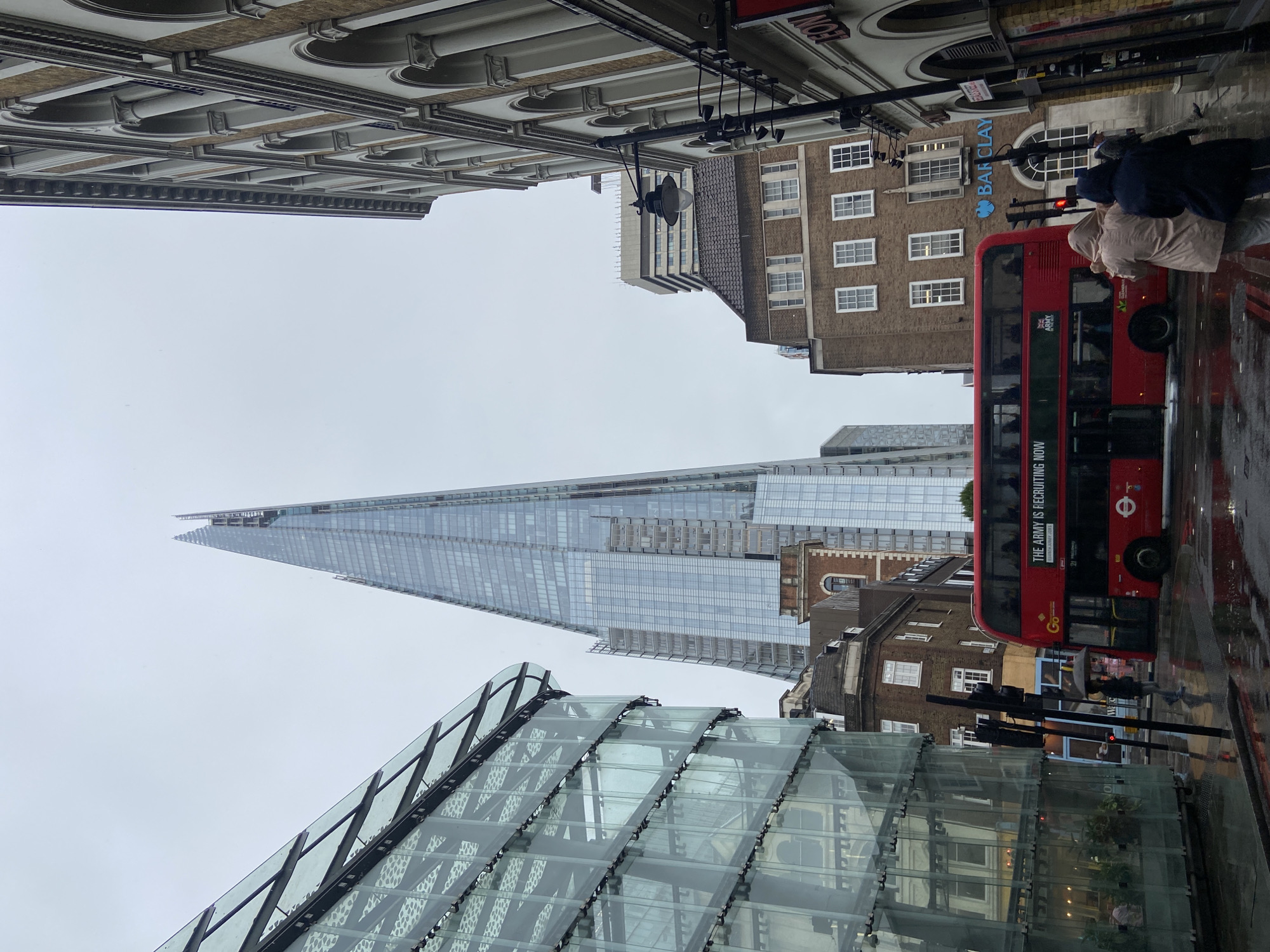 This vertically oriented image captures a bustling street scene in London on an overcast and wet day. The most prominent subject is The Shard, a tall, slender glass skyscraper, dominating the center of the frame against a grey sky. In the foreground, to the bottom left, is a contemporary glass-paneled structure with geometric patterns.
On the right side of the street, a bright red London double-decker bus is visible, seemingly stopped or moving slowly, with reflections of the city appearing on its windows. Several people are on the pavement; two individuals are walking near the bus, and a person further down the street holds an umbrella.
Architecture varies from modern glass structures to older, traditional buildings. An older brick building with numerous white-framed windows on the right features a prominent blue "Barclays" bank logo. Overhead, an ornate building facade with arches and cornices is visible in the top left corner. The wet street surface, reflecting buildings and lights, indicates recent or ongoing rain, contributing to the subdued, overcast atmosphere.
Visible text includes: the "Barclays" logo on the bank building, "THE ARMY IS RECRUITING NOW" on a green advertisement panel on the side of the bus, and the circular "TfL" (Transport for London) logo also on the bus. A partially visible word "Go..." is on the bus. The scene portrays a characteristic urban environment of London during daytime with typical British weather.