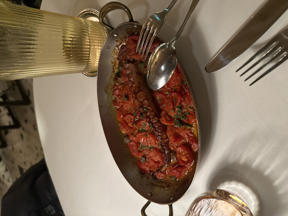 A silver pan filled with tomatoes and octopus rests on a white tablecloth, accompanied by a fork, spoon, knife, and a water glass. The dish is garnished with fresh herbs. A ridged glass pitcher sits nearby. A dark jacket is visible in the lower left corner, indicating a person is present.