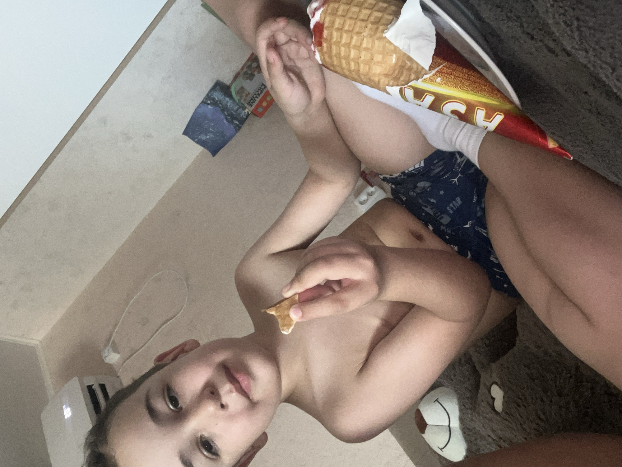 The image shows a young boy with a piece of waffle in his hand. He is shirtless and wearing dark blue shorts with a white star pattern. He is looking towards the camera with his mouth slightly open. Behind him, a part of an ice cream cone wrapper is visible, with Russian text that appears to read "АСА". A stuffed bear is also visible in the lower right corner of the image. The background consists of a light-colored tiled wall and floor. The scene appears to be indoors, possibly in a home setting. There are no clear indications of the time of day or weather. The boy's expression is neutral.