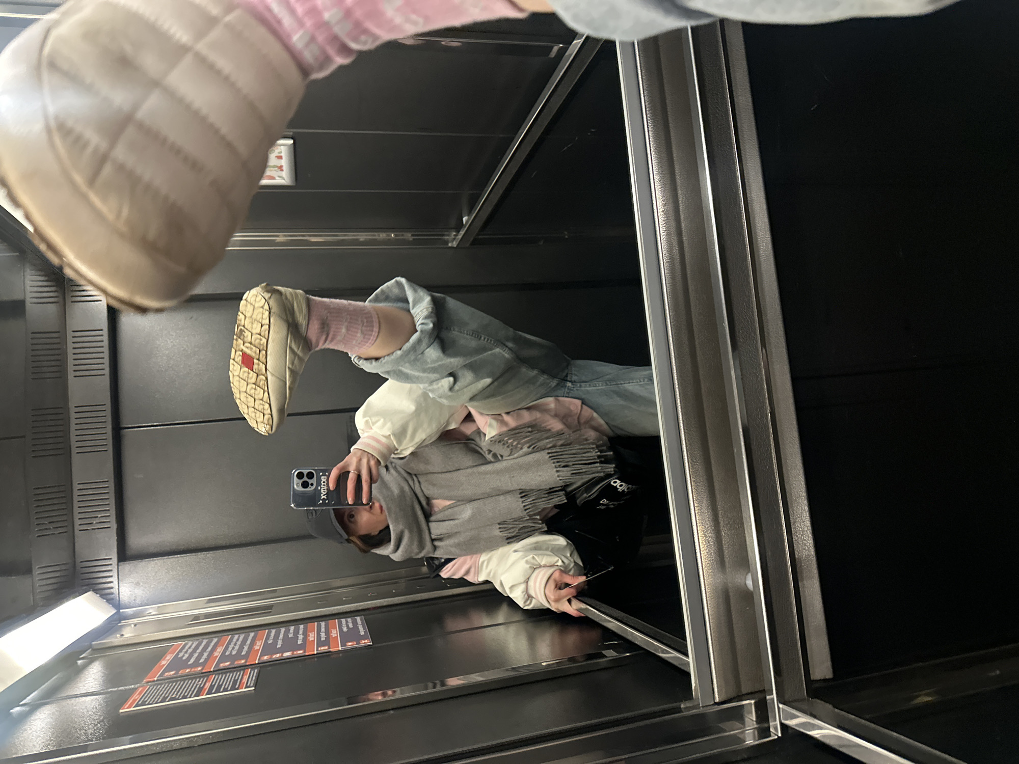 The image, rotated 90 degrees clockwise, depicts a person taking a mirror selfie inside what appears to be an elevator in Perm, Russia. The perspective is from the person holding the camera, looking upwards towards a highly reflective ceiling or upper wall, capturing their own reflection.
The primary subject is the reflection of a person, seen horizontally due to the image's rotation. They are dressed in warm, casual attire suitable for a cold climate. This includes a light-colored, possibly white or cream, puffy jacket with pink striped cuffs and a pink interior, light blue jeans, and pink socks. A grey scarf with fringed ends is wrapped around their neck. They are wearing a dark cap and light-colored, sturdy winter boots. One of these boots is prominently featured in the top left foreground, appearing close to the camera lens, indicating it belongs to the person taking the photo.
The person is holding a dark-colored smartphone, likely an iPhone given the camera module design, which is pointed towards the mirrored surface to capture the selfie. A black bag, possibly with a faint "Adidas" logo visible in the reflection, is resting near the person's feet in the reflection.
The setting is an elevator interior, characterized by dark, possibly black, walls and bright, metallic silver trim framing the panels and corners. Bright light sources are visible, illuminating the reflective surfaces. On one of the reflected walls, several rectangular signs or stickers with red and white text are affixed, but the text content is illegible due to the reflection and resolution.
The main activity is a mirror selfie, capturing the person's outfit and appearance. The person's face is partially visible in the reflection.
Notable details include the casual winter fashion, the specific camera angle, and the high reflectivity of the elevator surfaces. The smartphone case features reversed white text that reads "BODTAX". The overall impression is a personal moment captured in a public, functional space.